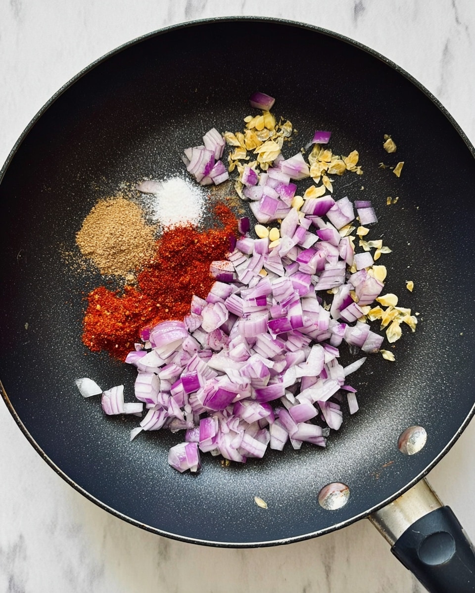 A top view of a black frying pan on a white marbled surface showing chopped purple and white onion pieces spread mostly on the right side, with small yellow chopped garlic pieces over them. On the left side of the pan, there are three piles of ground spices arranged close together: a light brown powder, a white powder, and a vibrant red powder. The pan handle is visible on the bottom right. photo taken with an iphone --ar 4:5 --v 7