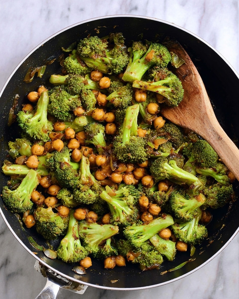 A black frying pan filled with cooked broccoli and chickpeas sits on a white marbled surface, with a wooden spatula resting inside the pan. The broccoli florets are bright green with some parts slightly browned, and the chickpeas are golden brown, mixed evenly throughout the pan. The dish has a glossy texture from a light sauce coating the vegetables. Photo taken with an iphone --ar 4:5 --v 7