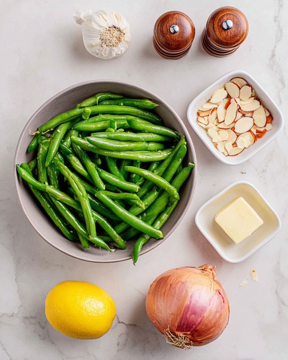 The image shows raw green beans in a gray bowl at the top left, surrounded by other ingredients on a white marbled surface. To the top right is a small white bowl with two square slices of yellow butter. Below the butter is a bright yellow lemon half, centered slightly right of the image. Beneath the lemon is a white bowl filled with chopped garlic. To the bottom right is a white bowl containing sliced almonds with brown edges. Near the center left, there is a whole shallot with a pinkish-brown skin. At the bottom left are two brown wooden salt and pepper shakers marked with