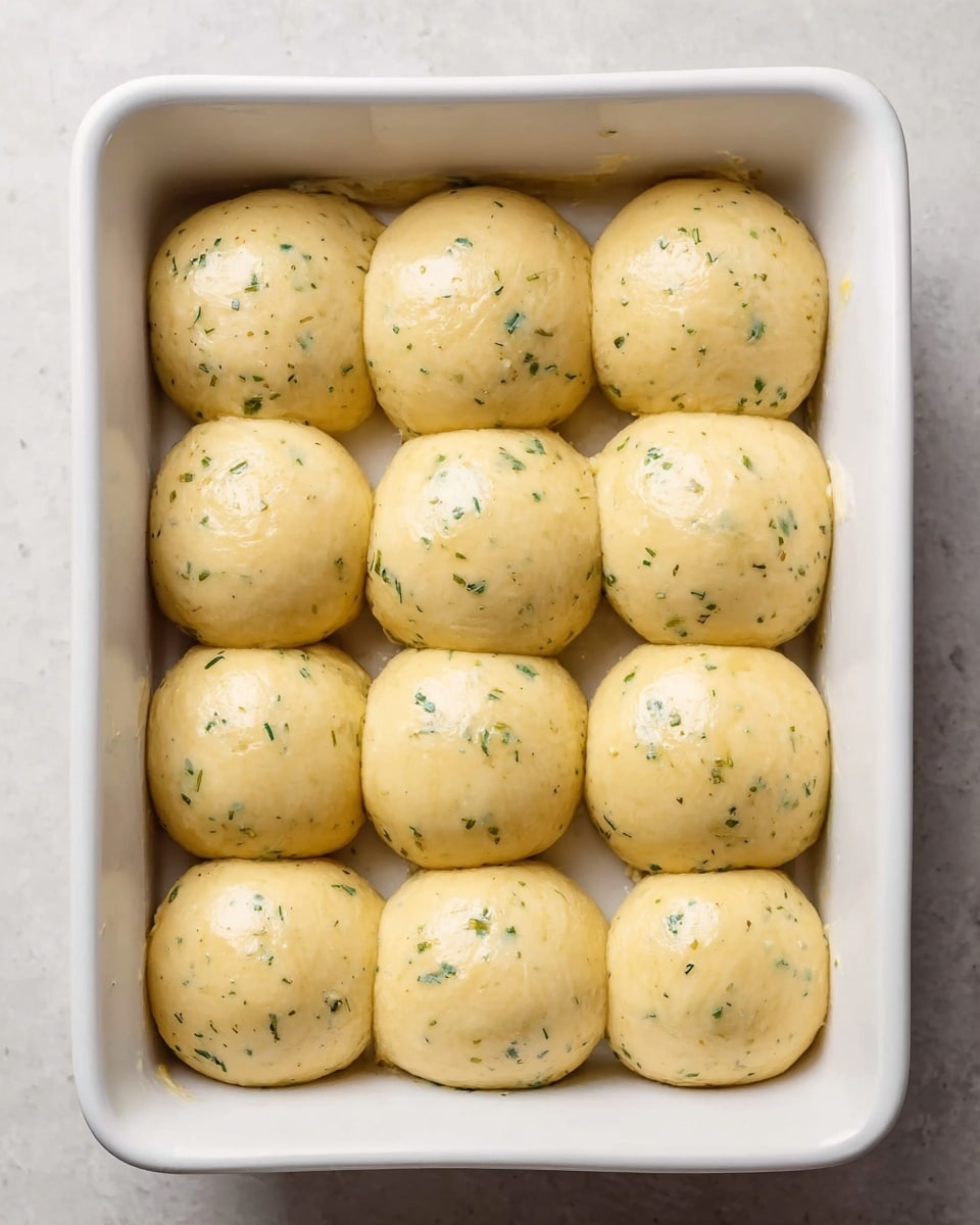 A white rectangular baking dish holds twelve smooth, round dough balls arranged in a 3x4 grid. Each dough ball has a glossy, light golden-yellow surface speckled with small green herb bits, giving a fresh and slightly textured look. The dough balls are touching each other gently, filling the dish evenly. The dish sits on a light white marbled surface. photo taken with an iphone --ar 4:5 --v 7