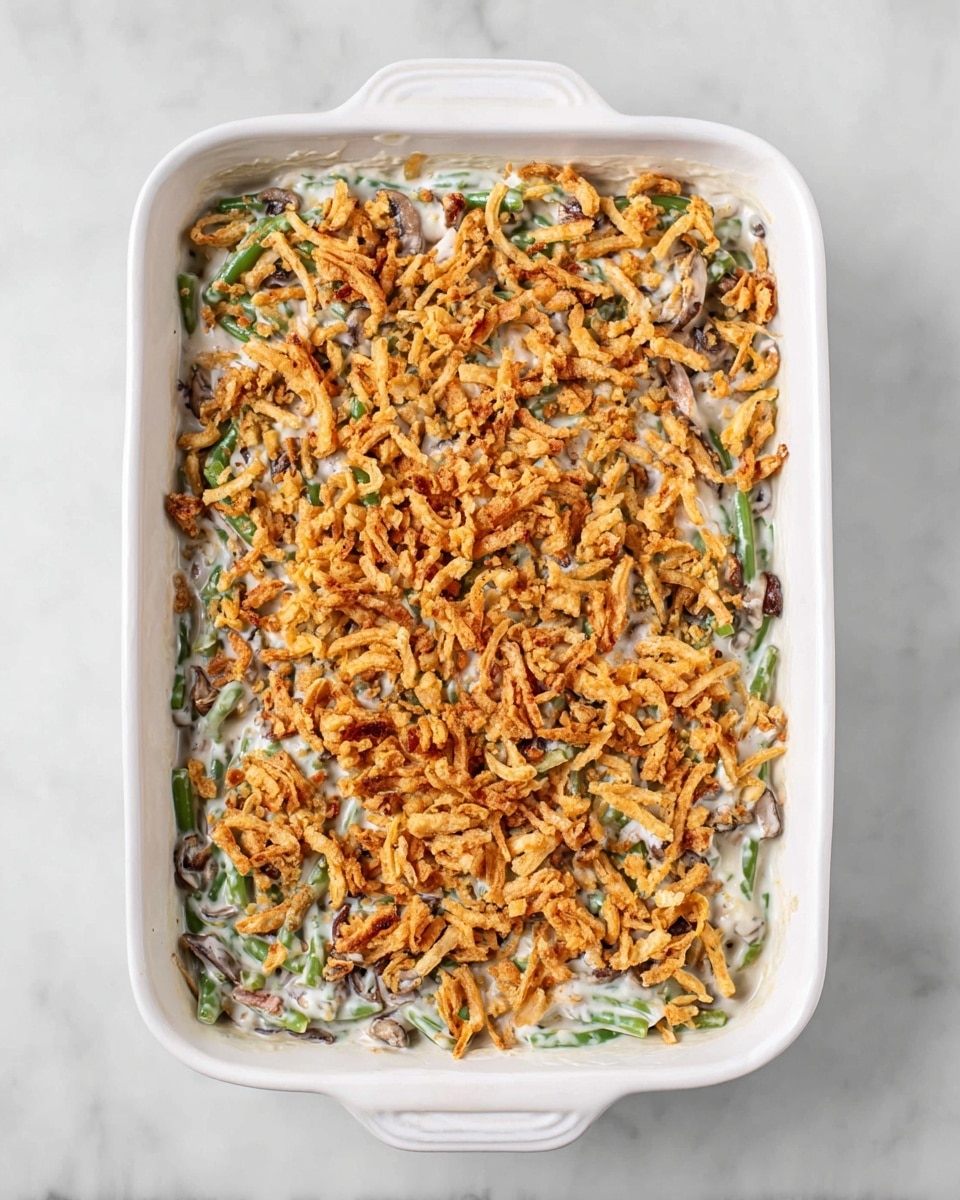 The image shows a white rectangular baking dish filled with a layered green bean casserole. The bottom layer contains chopped green beans mixed with creamy white sauce and small bits of browned mushrooms and onions, creating a soft and mixed texture. The top layer is evenly covered with golden crunchy fried onions scattered all over, adding a crispy contrast to the creamy green beans underneath. The dish sits on a white marbled surface, and the photo is taken from above, capturing the full view of the casserole photo taken with an iphone --ar 4:5 --v 7