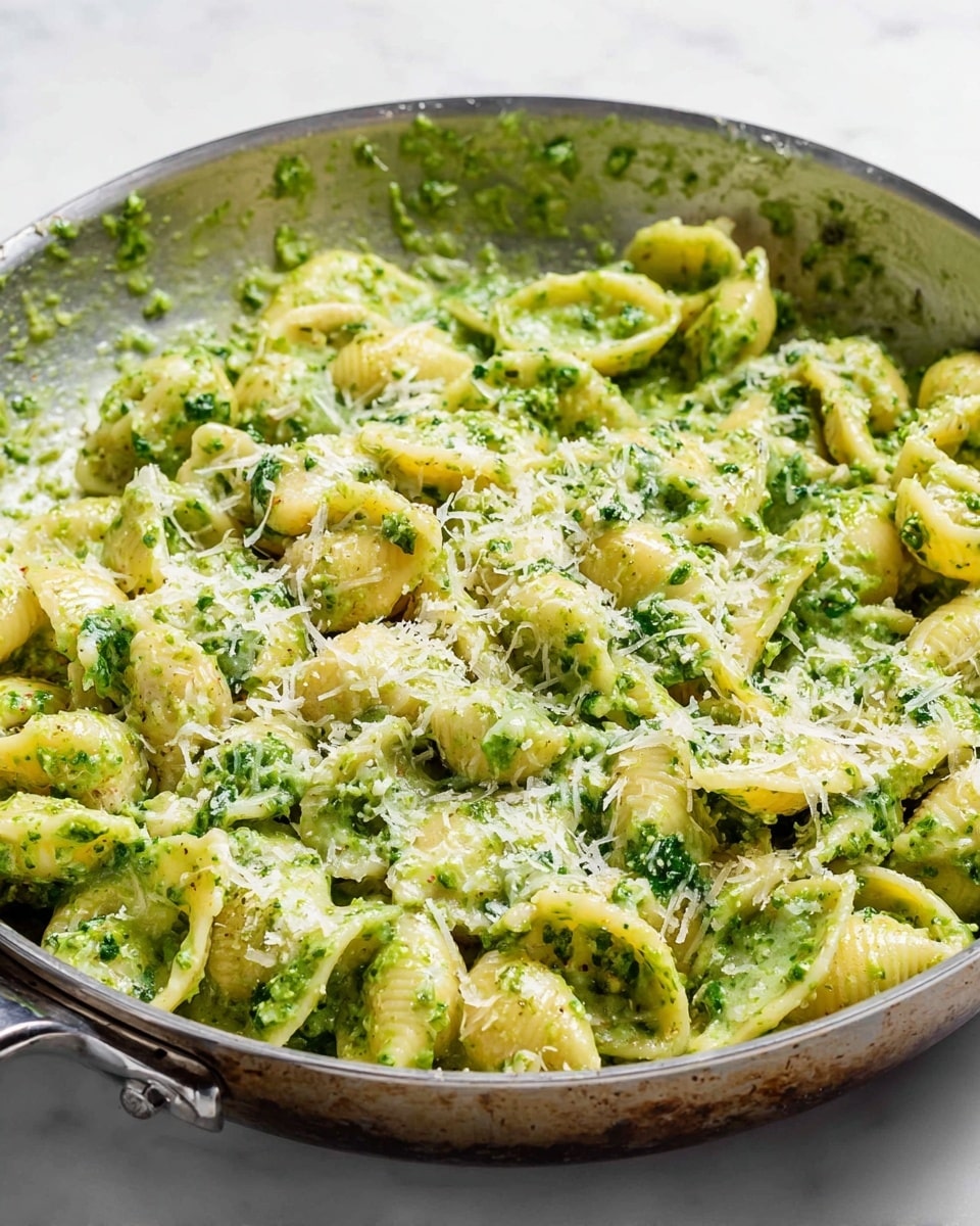A silver pan holds a creamy pasta dish with about two layers of small shell-shaped noodles richly coated in a vibrant green pesto sauce that looks smooth yet slightly textured with bits of herb. On top, a light sprinkle of finely grated pale yellow cheese adds a delicate contrast, scattered evenly. The pan shows reflections of light on its worn metallic surface and rests on a white marbled texture background. Photo taken with an iphone --ar 4:5 --v 7
