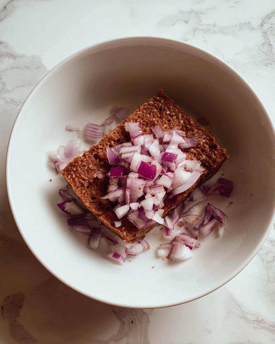 A white bowl sits on a white marbled surface, inside it is a square piece of brown textured food that looks soft and moist. On top of and around the brown piece, there are small chopped pieces of red onion with purple skins and white insides spread unevenly. The scene is lit softly, showing clear details and colors. photo taken with an iphone --ar 4:5 --v 7
