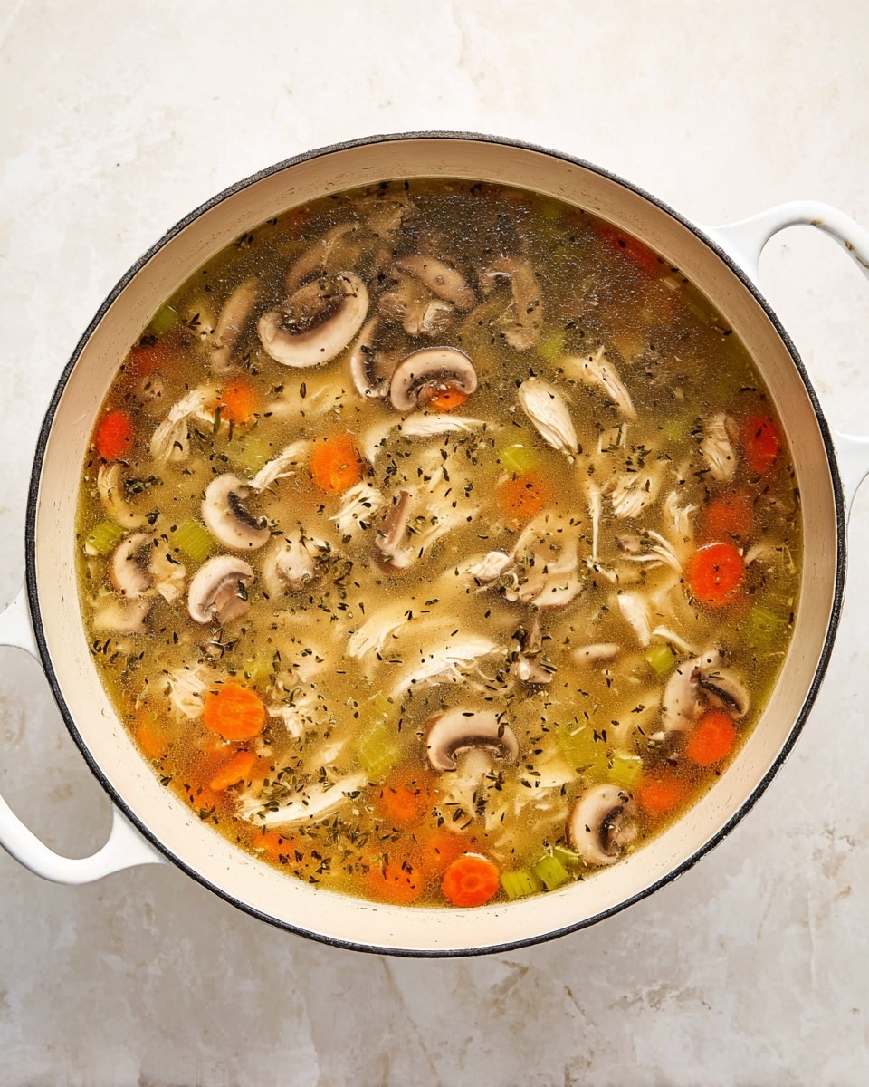 A light golden broth fills a large white pot, layered with thinly sliced white and brown mushrooms floating on top, mixed with small bright orange carrot cubes and round green celery slices. Shredded white pieces of chicken are scattered throughout the clear soup, with tiny dark green herb flakes sprinkled over everything. The pot has white handles and is set on a white marbled surface. photo taken with an iphone --ar 4:5 --v 7