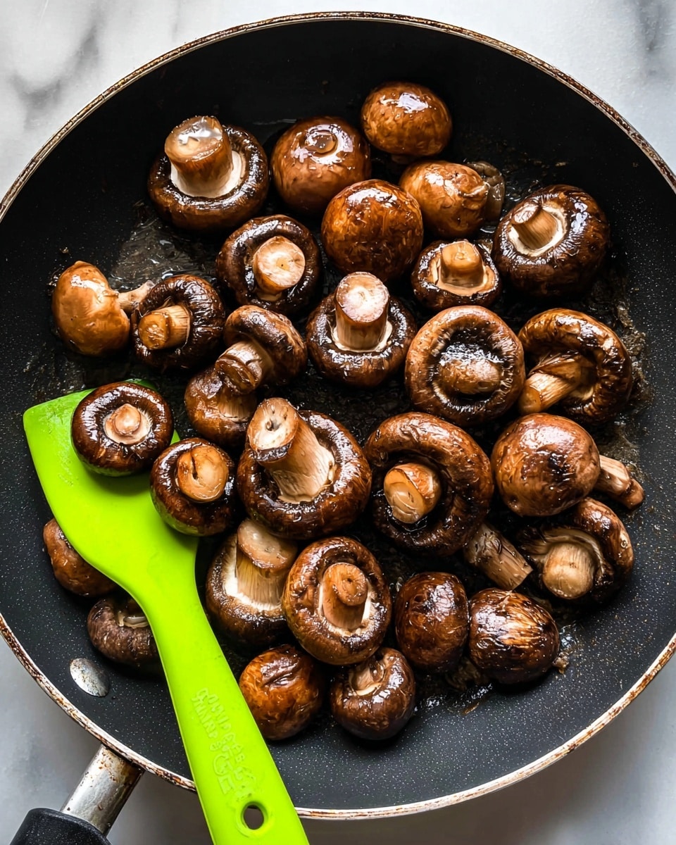 A black frying pan filled with about three dozen cooked whole brown mushrooms, with their caps shiny and slightly wrinkled from cooking. The mushrooms vary slightly in size and are spread unevenly across the pan, some with stems facing up and others turned sideways, showing their textured undersides. On the left side of the pan, a bright green silicone spatula rests partially under a cluster of mushrooms. The pan sits on a white marbled surface that contrasts with the dark frying pan and the earth tones of the mushrooms, photo taken with an iphone --ar 4:5 --v 7