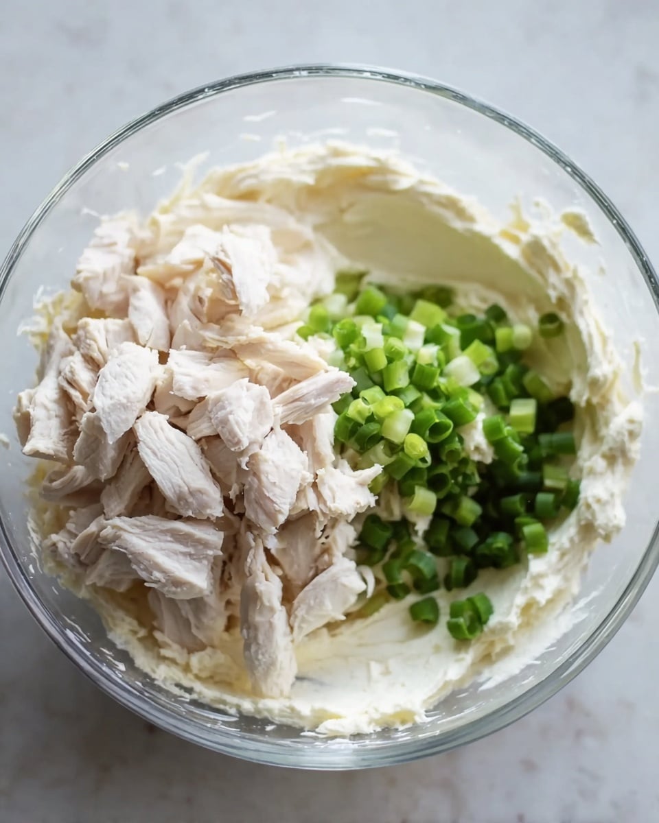 A clear glass bowl sits on a white marbled surface holding three main visible layers of ingredients: a base layer of thick, creamy white mixture spread around the bowl, a middle pile of small, pale beige chunks of cooked chicken stacked mostly on one side, and a cluster of chopped bright green onions placed next to the chicken. The textures range from smooth and creamy to firm and meaty next to the fresh green topping. The scene looks bright with soft natural light, showing the details and colors clearly. Photo taken with an iphone --ar 4:5 --v 7