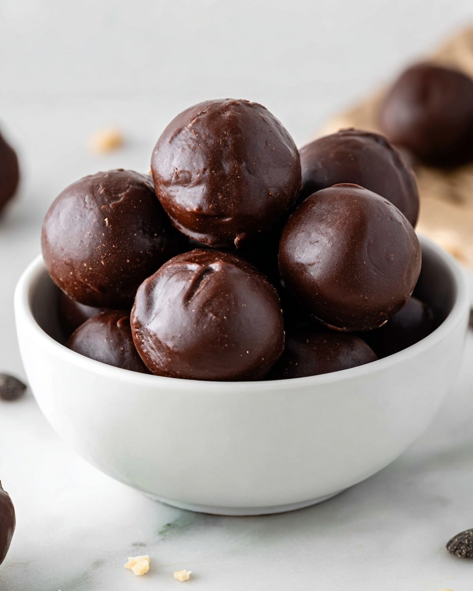 A white bowl filled with round chocolate-covered treats stacked in a small pile, each piece showing a smooth, shiny dark brown chocolate coating that has slight texture and natural swirls on the surface, giving them a rich and dense look. The bowl sits on a white marbled surface with a few small crumbs scattered nearby, and the background is softly blurred to focus on the chocolate balls. photo taken with an iphone --ar 4:5 --v 7