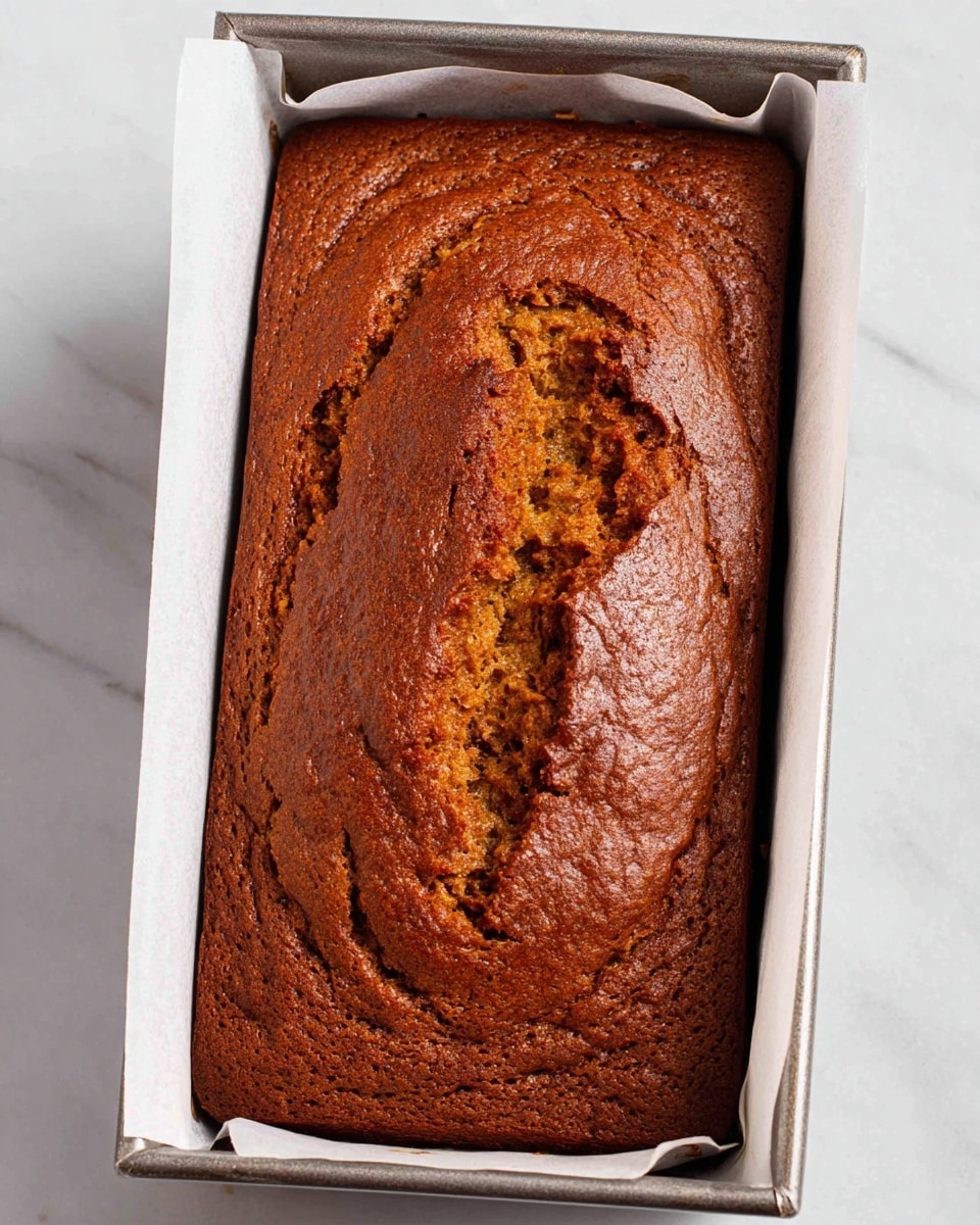 A single rectangular loaf cake with a warm brown crust sits inside a metal baking pan lined with white parchment paper. The cake has a cracked, textured top layer showcasing a slightly darker, moist interior beneath the surface. The edges are evenly browned and the cake's top has some uneven raised areas, giving it a rustic look. The pan rests on a white marbled surface. photo taken with an iphone --ar 4:5 --v 7