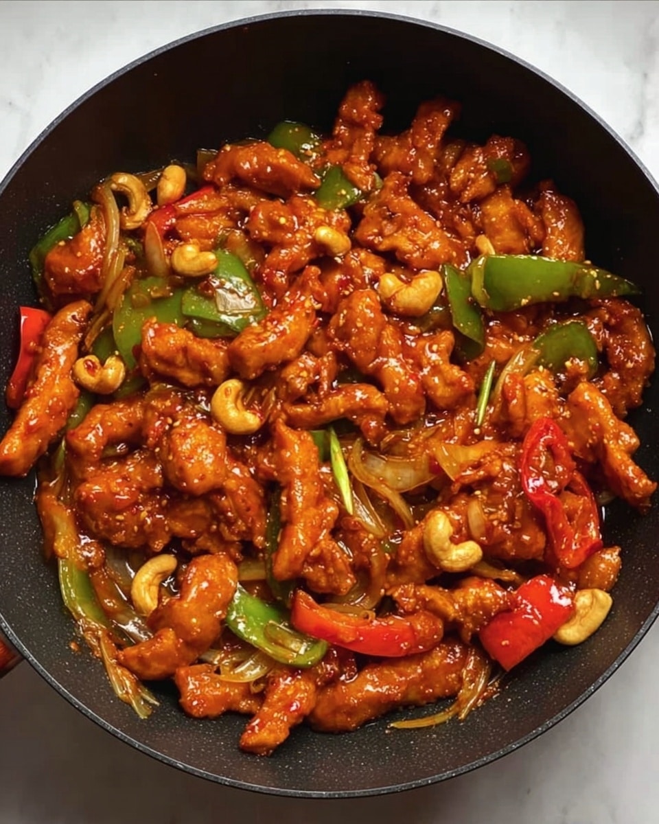 The image shows a dark round pan filled with many pieces of glossy, sauce-coated chicken strips in a rich orange hue, mixed with sliced green and red bell peppers, whole cashew nuts, and thin translucent onion slices. The chicken strips have a slightly crispy texture, and the vegetables add bright spots of green and red around the pan. The sauce looks thick and shiny, covering all ingredients evenly, giving a vibrant and appetizing look. The pan sits on a white marbled surface. photo taken with an iphone --ar 4:5 --v 7