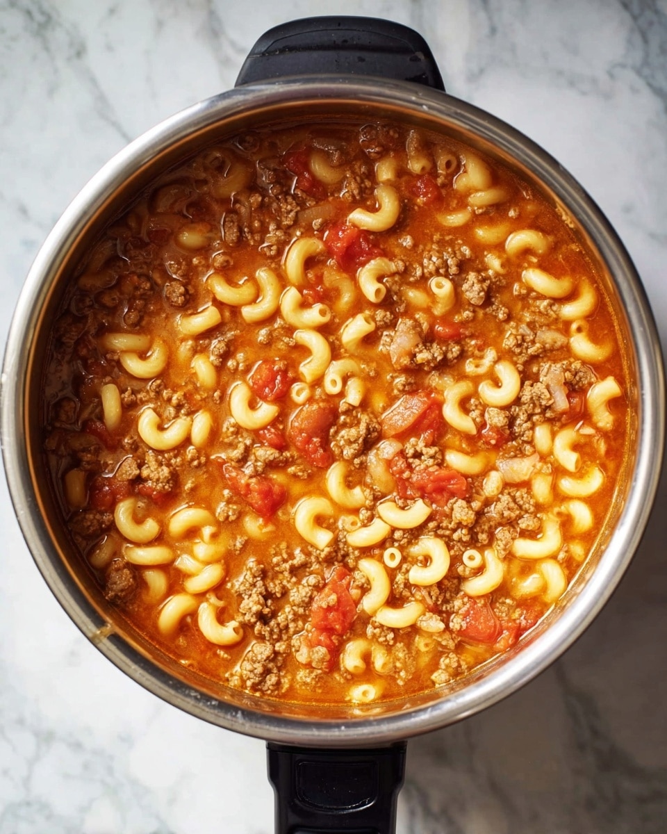 A top view of a stainless steel pot filled with a simmering mixture of elbow macaroni, browned ground meat, and chunky pieces of red tomato in a thin, orange-red broth, all evenly spread inside the pot. The pot is sitting on a surface with a white marbled texture. photo taken with an iphone --ar 4:5 --v 7