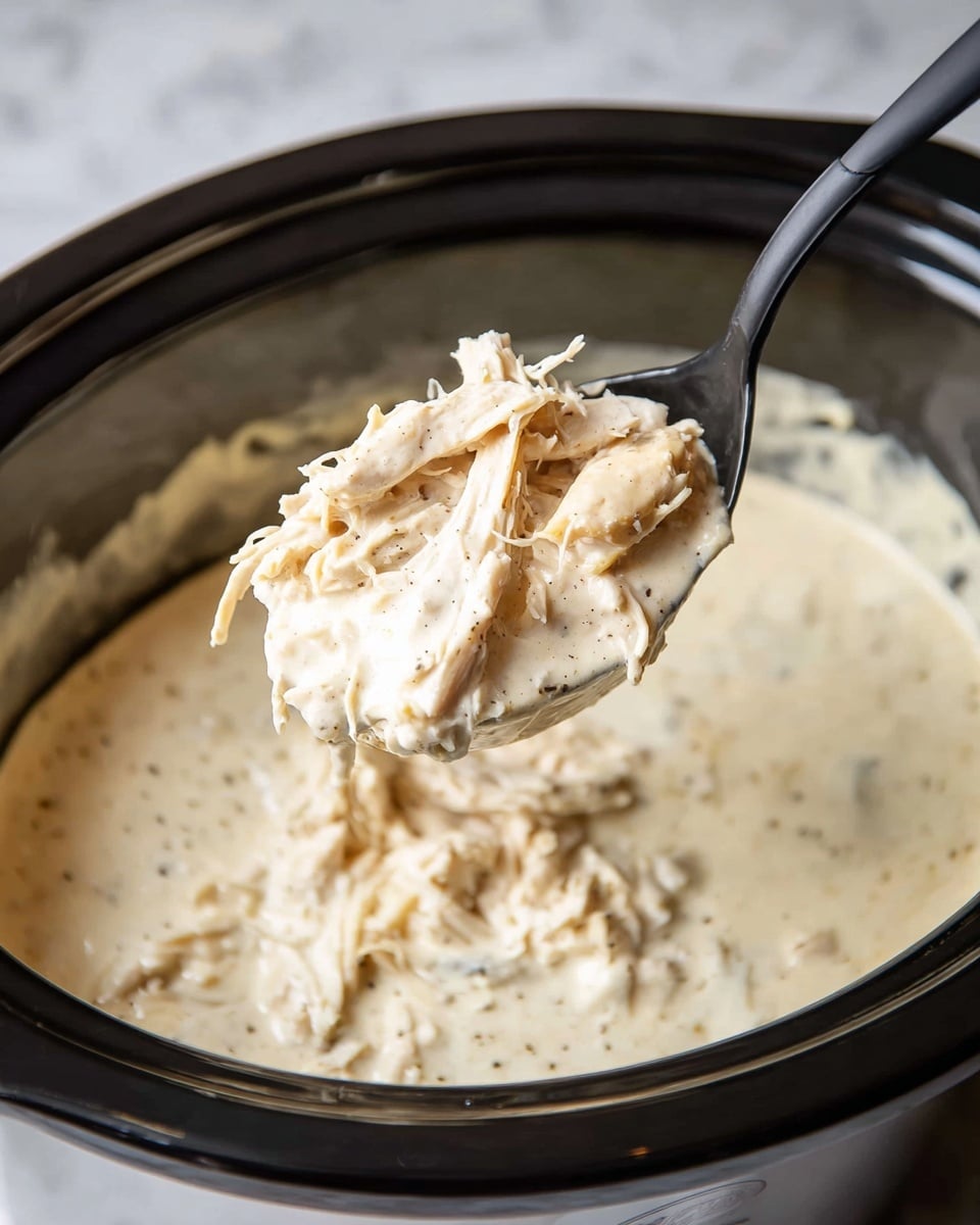 The image shows a black slow cooker filled with a thick creamy white sauce mixed with shredded light beige chicken pieces. A black spoon lifts a scoop of the mixture from the cooker, showing soft shredded chicken layered in a smooth, pale cream sauce with small specks of black pepper. The slow cooker sits on a white marbled surface. photo taken with an iphone --ar 4:5 --v 7