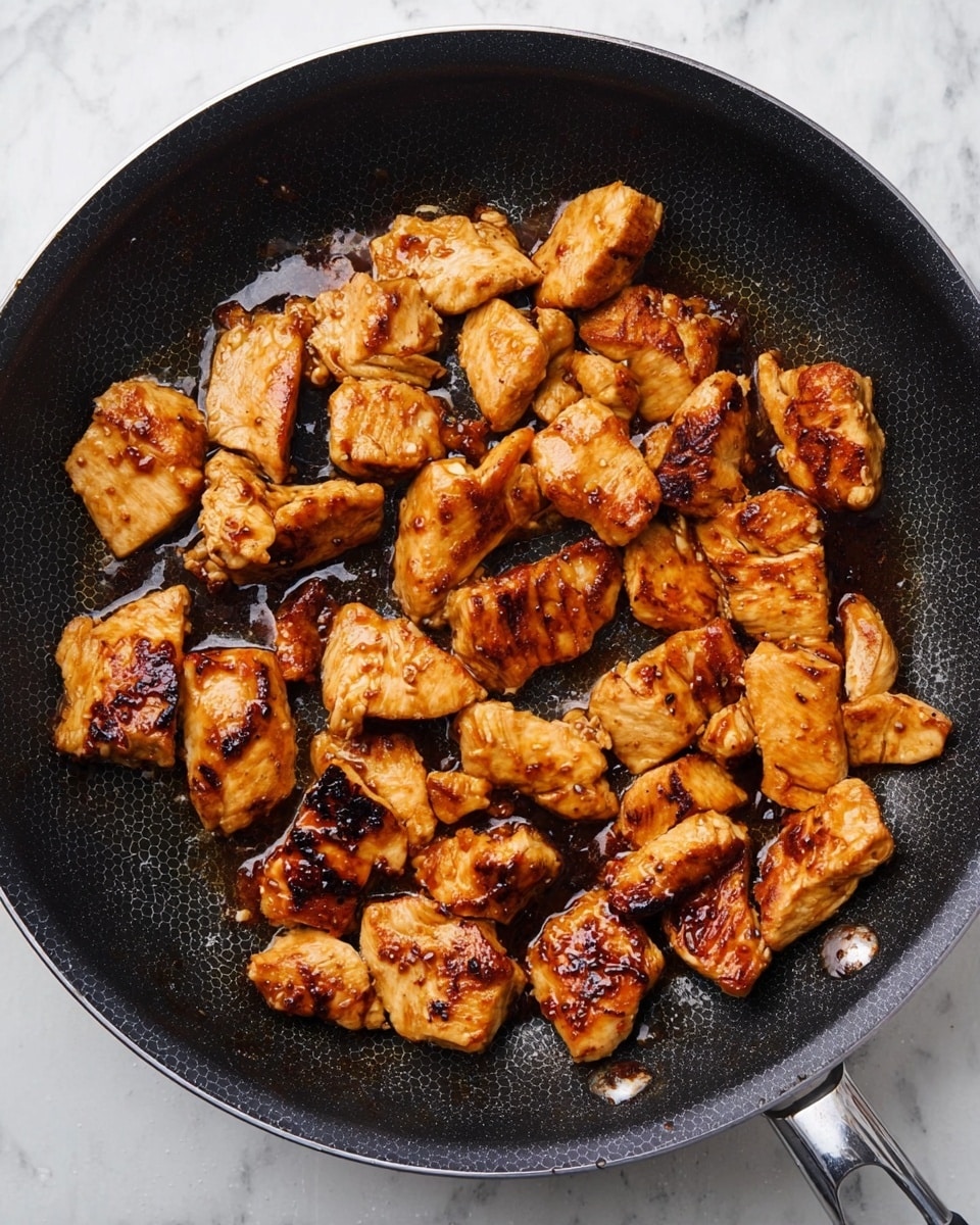 The image shows a black frying pan filled with several pieces of cooked chicken. The chicken pieces are golden brown with darker grill marks and a shiny sauce coating. The pieces vary in size and shape and are spread out evenly in the pan, which has a hexagonal pattern visible under the food. The pan rests on a white marbled surface, creating a clean and bright background. photo taken with an iphone --ar 4:5 --v 7