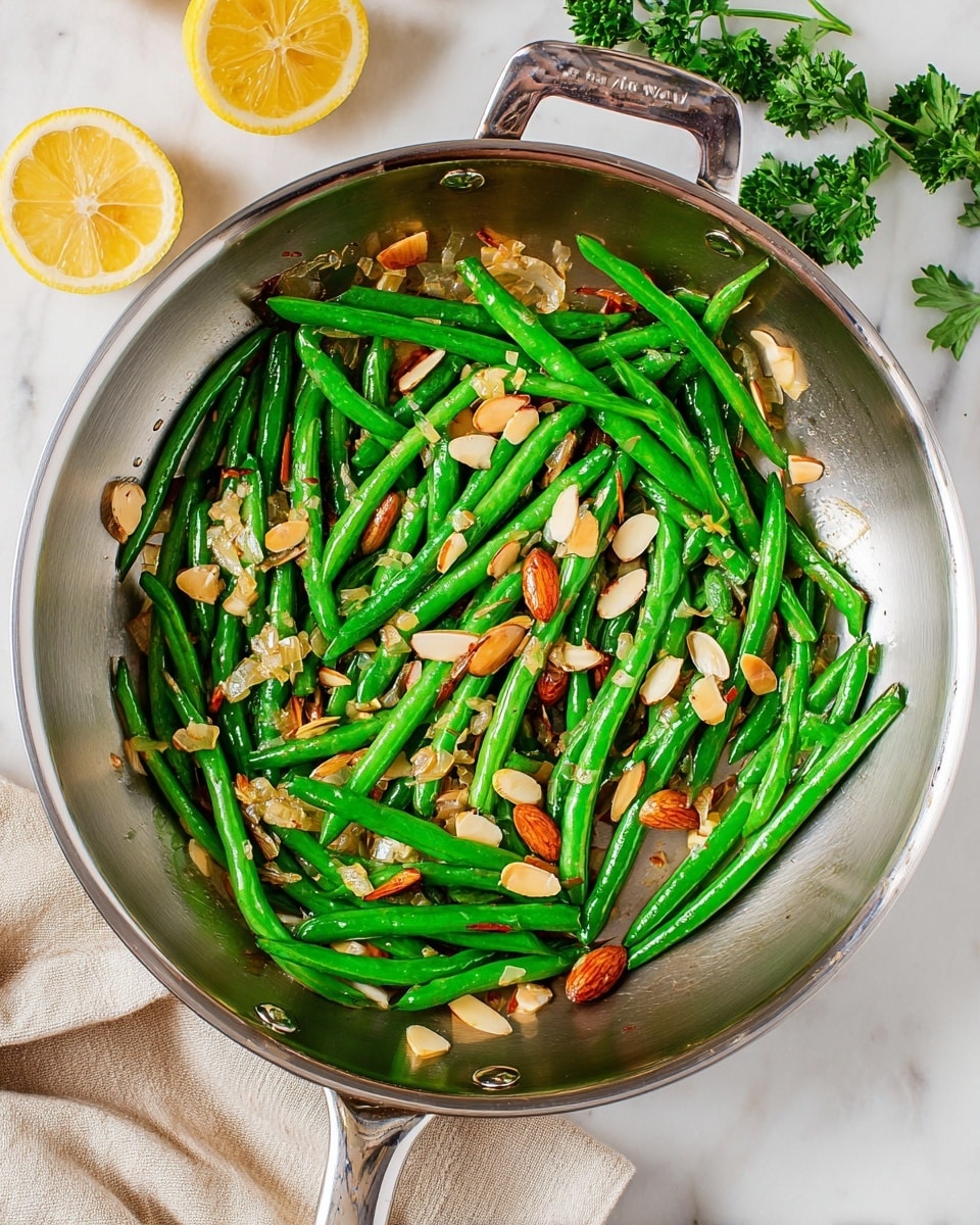A shiny silver pan holds bright green cooked green beans mixed with light golden brown sliced almonds and small bits of translucent cooked onion, all slightly oily giving a fresh, glistening look; the pan has a sturdy silver handle and rests on a white marbled surface with a few green parsley leaves and bright yellow lemon slices nearby. photo taken with an iphone --ar 4:5 --v 7