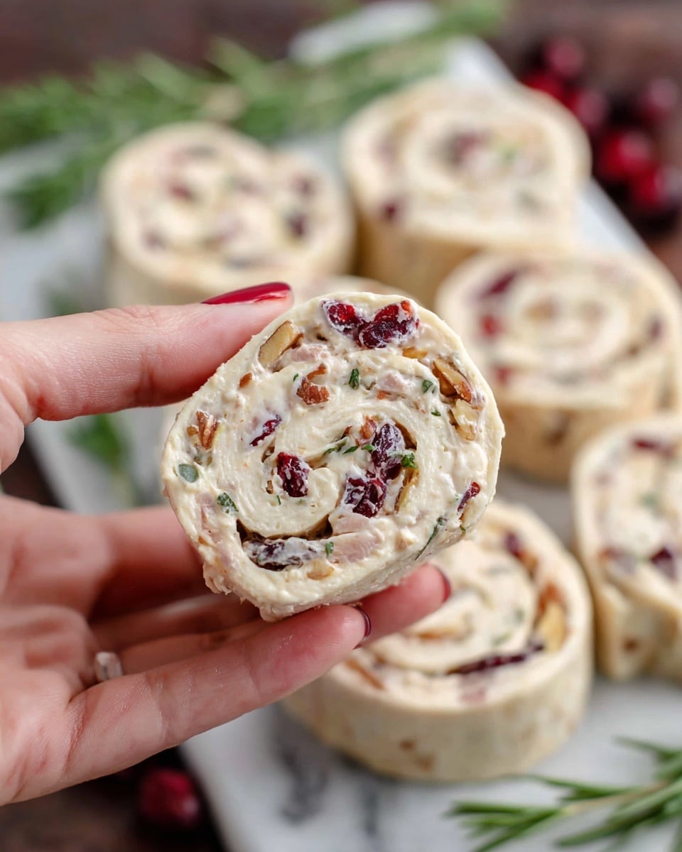 A woman's hand is holding a white tortilla roll with visible layers of creamy filling mixed with small pieces of red dried cranberries and bits of nuts. The roll is sliced into thick pinwheel shapes, showing a spiral pattern inside. Behind the hand, more similar pinwheel slices are arranged on a white marbled surface, with a few sprigs of green herbs for decoration. The overall colors are light beige from the tortilla, white from the filling, and pops of red and brown from the cranberries and nuts. Photo taken with an iphone --ar 4:5 --v 7