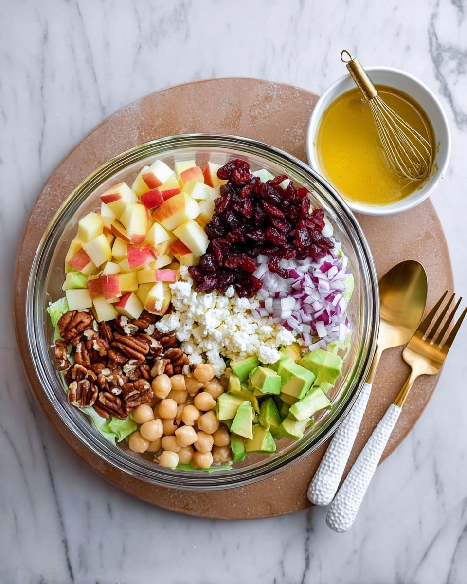 A clear glass bowl resting on a white marbled board contains seven different layers of salad ingredients. Starting from the top left side is a layer of yellow and red chopped apple pieces, next to it is a pile of deep red dried cranberries mixed with small pieces of purple onion. Below the cranberries is a clump of crumbly white cheese. To the bottom right of the bowl are bright green avocado cubes. Towards the bottom left sits a cluster of brown pecan halves, and above them are pale round chickpeas. A small white bowl with a golden yellow salad dressing and a small whisk is placed to the left of the bowl, all set on a white marbled surface. To the right of the bowl are a gold spoon and fork with white textured handles. photo taken with an iphone --ar 4:5 --v 7