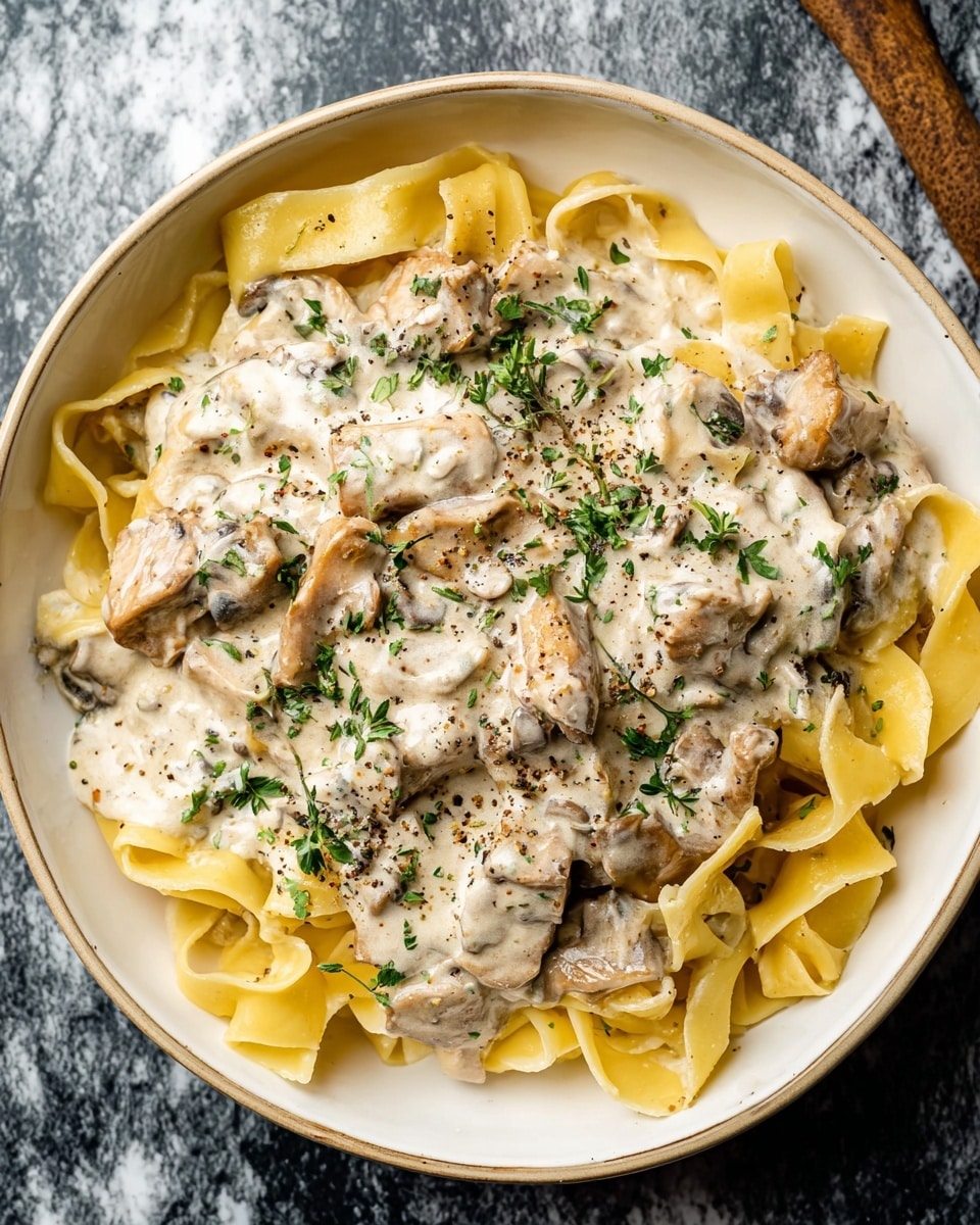 The image shows a creamy pasta dish inside a black pot. The pasta is light yellow, wide, and flat layered unevenly throughout the creamy white sauce. The sauce has a smooth texture with specks of black pepper. Brown mushroom slices and chunks of brown meat are mixed in, adding texture and color contrast. Small green herbs are sprinkled over the top for a fresh look. The sauce clings to the pasta and ingredients, creating a rich and cozy feel. photo taken with an iphone --ar 4:5 --v 7