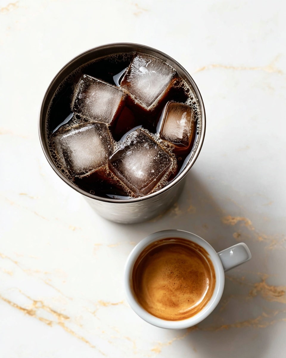 A tall metal cup filled with dark iced coffee contains five clear ice cubes floating on top, with light froth and small bubbles around the edges. Next to it, a small white espresso cup holds a little dark coffee with a smooth, golden crema layer on top. Both the cup and espresso rest on a white marbled surface with subtle gold veining, creating a clean and elegant look. Photo taken with an iphone --ar 4:5 --v 7