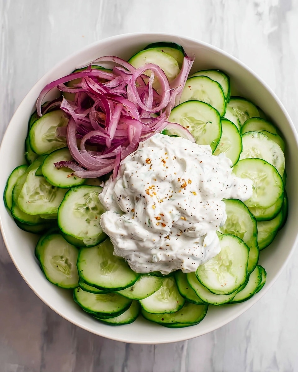 A white bowl filled with thinly sliced green cucumber arranged in a circular pattern covering most of the bowl's surface. On the left side, there is a small pile of thin, long, purple-red onion slices. On top in the center lies a dollop of creamy white sauce with visible small black and brown seasoning bits, having a thick and textured appearance. The bowl is placed on a white marbled surface. Photo taken with an iphone --ar 4:5 --v 7