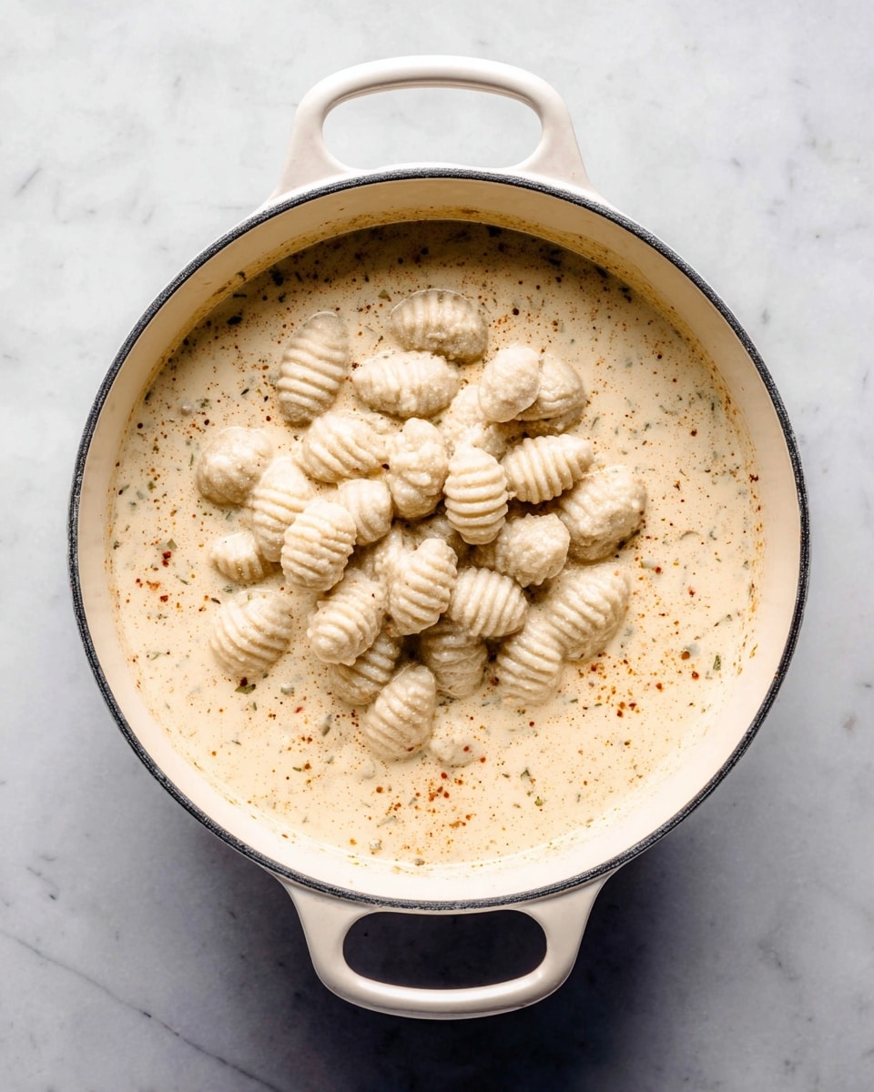 A white pot with two handles is filled with a creamy, light beige sauce that has small specks of spices and herbs throughout. On top of the sauce, there is a pile of small, pale gray gnocchi with ridged lines and smooth textures, sitting in the center of the pot. The pot is placed on a white marbled surface that adds a clean, bright background to the image. photo taken with an iphone --ar 4:5 --v 7