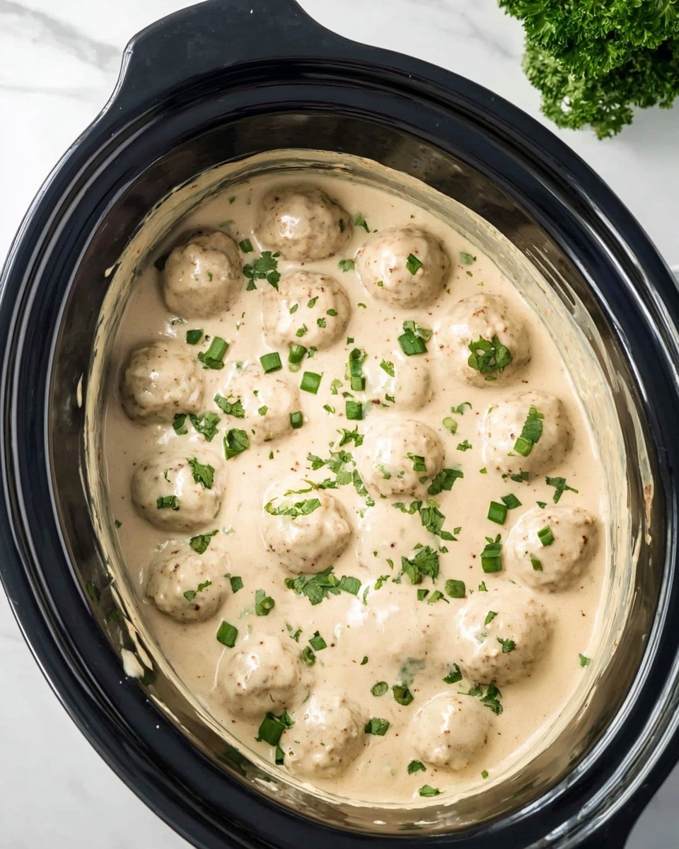 Inside a black slow cooker, there is a thick creamy sauce with a light beige color, covering round meatballs that are light brown and smooth. The sauce has a few specks of seasoning visible and is topped with small pieces of green chopped herbs scattered evenly across the surface. The slow cooker is placed on a white marbled surface with a small bunch of green herbs in the background. photo taken with an iphone --ar 4:5 --v 7