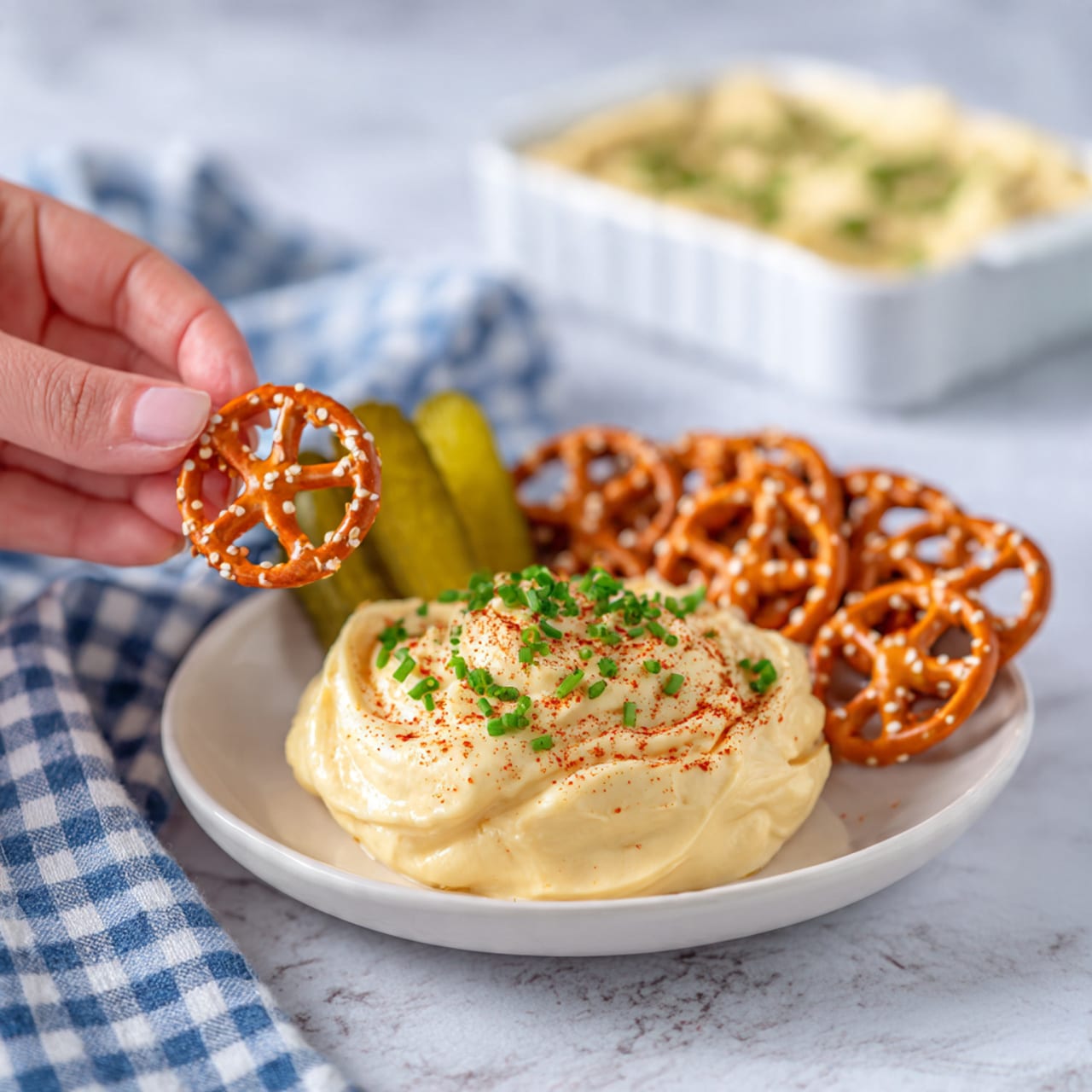A white plate on a white marbled surface holds a small pile of golden-brown pretzel crackers arranged in a slightly overlapping layer on the right side. Next to them on the left are two small green pickles. Behind the crackers is a large dollop of pale creamy yellow hummus sprinkled with tiny green chive pieces and a bit of red paprika on top. The hummus looks smooth with some soft ripples and is slightly bigger than the pickles. In the background, there is a white rectangular dish with more hummus inside, resting on a blue and white checkered cloth. photo taken with an iphone --ar 4:5 --v 7