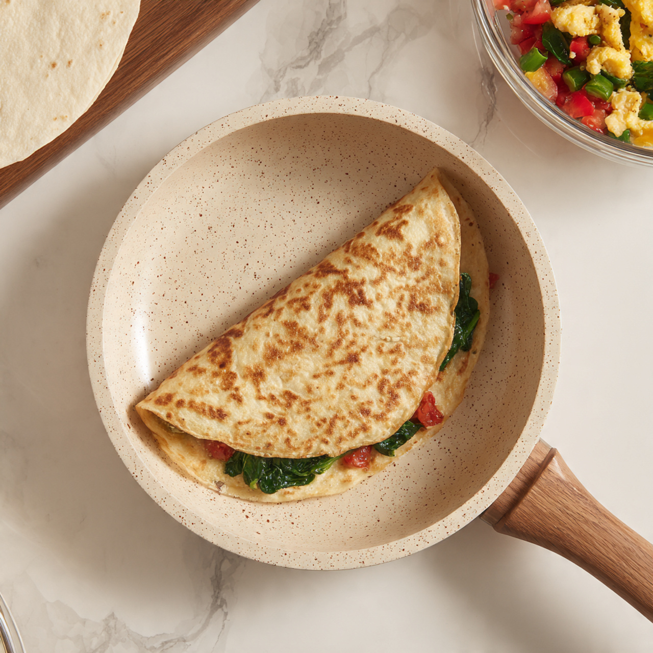 A folded tortilla with a warm, golden-brown toasted surface sits in a beige pan with tiny dark speckles and a wooden handle. The tortilla is folded in half, showing a peek of its filling: green leafy spinach and red tomato pieces inside. Nearby, a white flour tortilla rests on a white marbled surface to the left, and a clear glass bowl with a colorful mixture of chopped spinach, tomatoes, and scrambled eggs is partially visible to the upper right. The scene is bright and clean with a clear focus on the tortilla in the pan photo taken with an iphone --ar 4:5 --v 7