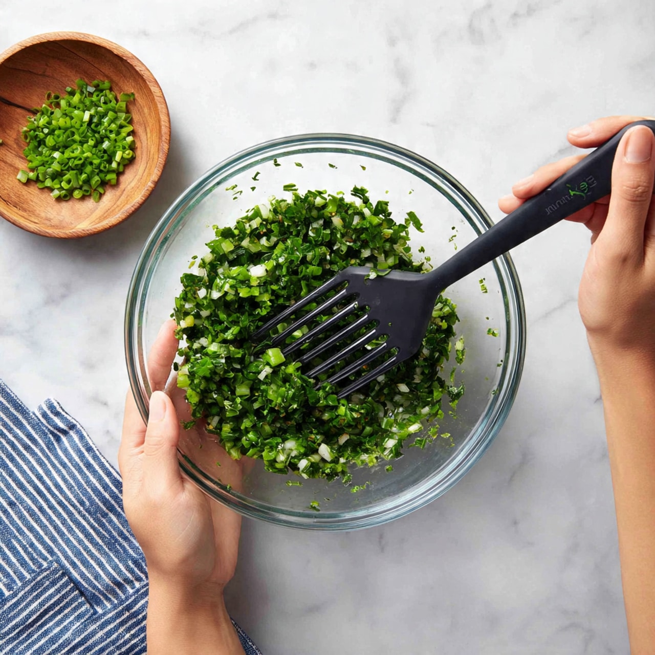 A clear glass bowl holds a thick, creamy white mixture with small chunks visible throughout. The bowl is held by a woman's left hand at the base, while a woman's right hand stirs the mixture using a black spatula with a white flexible edge. The bowl is placed on a white marbled surface with a small wooden bowl filled with chopped green chives in the upper left corner. Part of a blue striped cloth is visible near the bottom left edge of the image. photo taken with an iphone --ar 4:5 --v 7