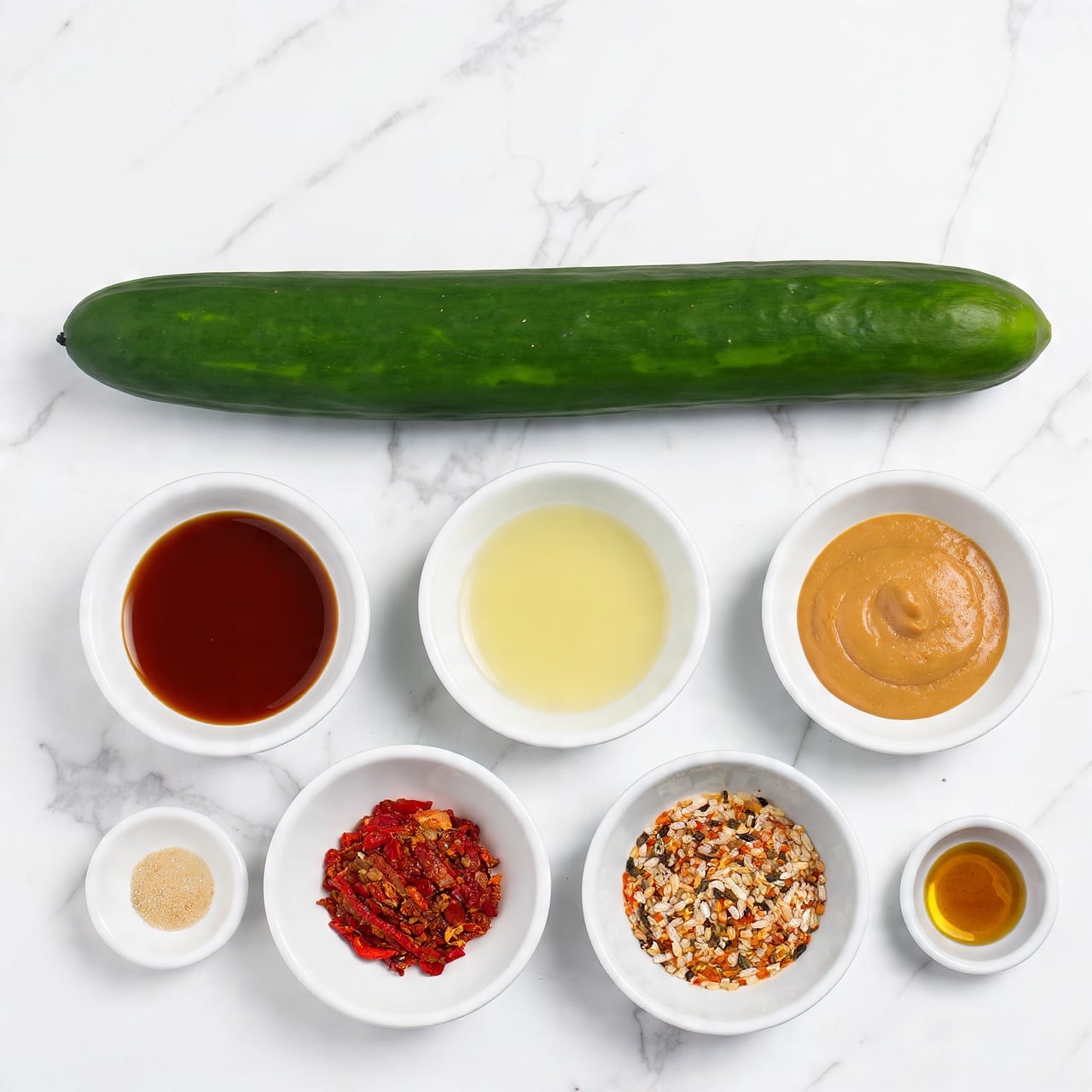 A long, dark green seedless cucumber lies at the top of the image against a white marbled texture background. Below it are seven small white bowls arranged in a loose circular pattern, each containing a different ingredient: a deep brown liquid labeled tamari or soy sauce, a pale yellow liquid labeled rice vinegar, a smooth, light tan runny peanut butter, a dark red chili crisp with visible chili flakes, a small pile of minced, light beige grated garlic, a heap of pale sesame seeds, and a small amount of dark amber toasted sesame oil. Each bowl's contents have smooth or grainy textures clearly visible, set neatly and cleanly. photo taken with an iphone --ar 4:5 --v 7