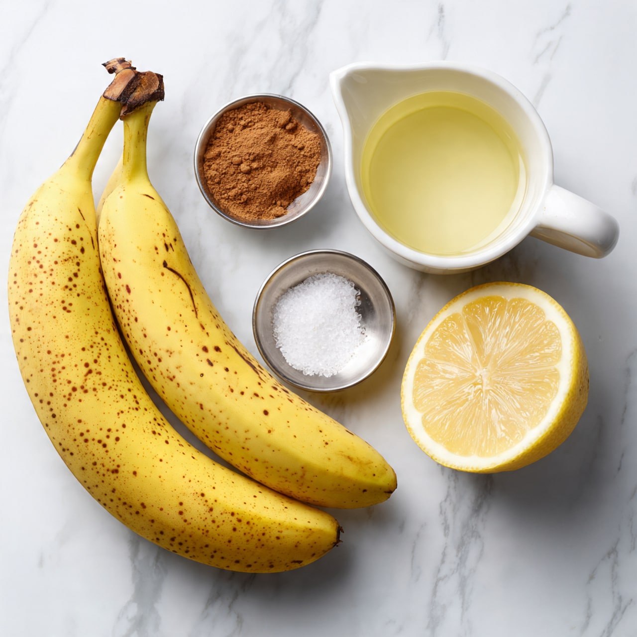 The image shows three yellow bananas with small brown spots lying together on a white marbled surface. To the right, there is a halved lemon showing its light yellow juicy inside. Below the lemon, there is a white small pitcher filled with clear coconut oil. Next to it are two small silver bowls; one contains a light brown cinnamon powder, and the other has white sea salt flakes. The items are neatly arranged and the overall setting is bright and clean, with a focus on natural colors and simple textures. photo taken with an iphone --ar 4:5 --v 7