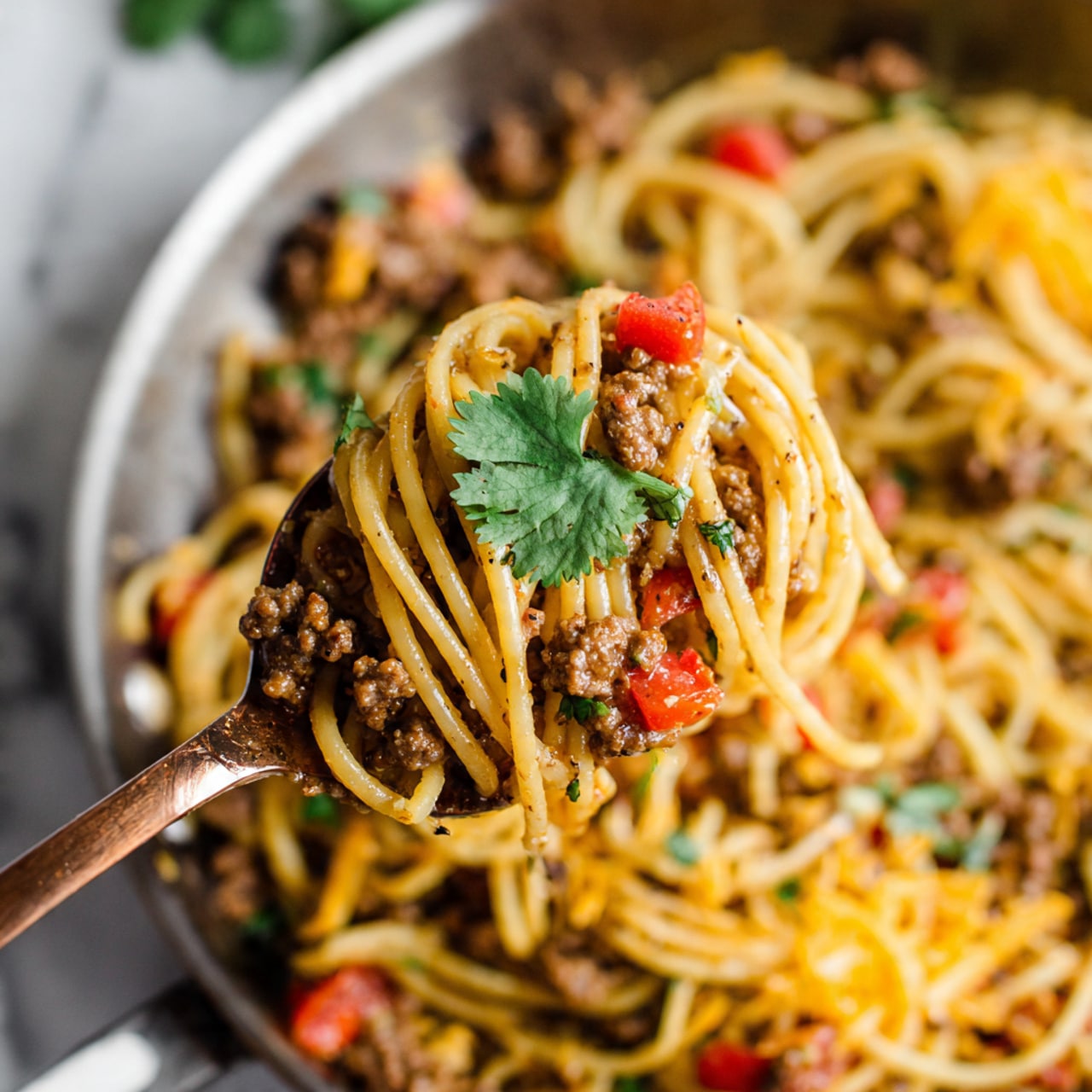 The image shows a close-up of a brass-colored spoon lifting a serving of spaghetti mixed with ground meat and diced red tomatoes. The spaghetti is golden yellow and slightly glossy, twisted around the spoon with visible black pepper specks. The meat is brown and crumbly, scattered evenly among the noodles, with bits of tomatoes adding bright red accents. A single green leaf of cilantro rests on top of the spaghetti on the spoon. The background features more spaghetti and meat mixture in a pan with a blurred block of yellow cheese and shredded cheese on a white marbled surface behind. Photo taken with an iphone --ar 4:5 --v 7