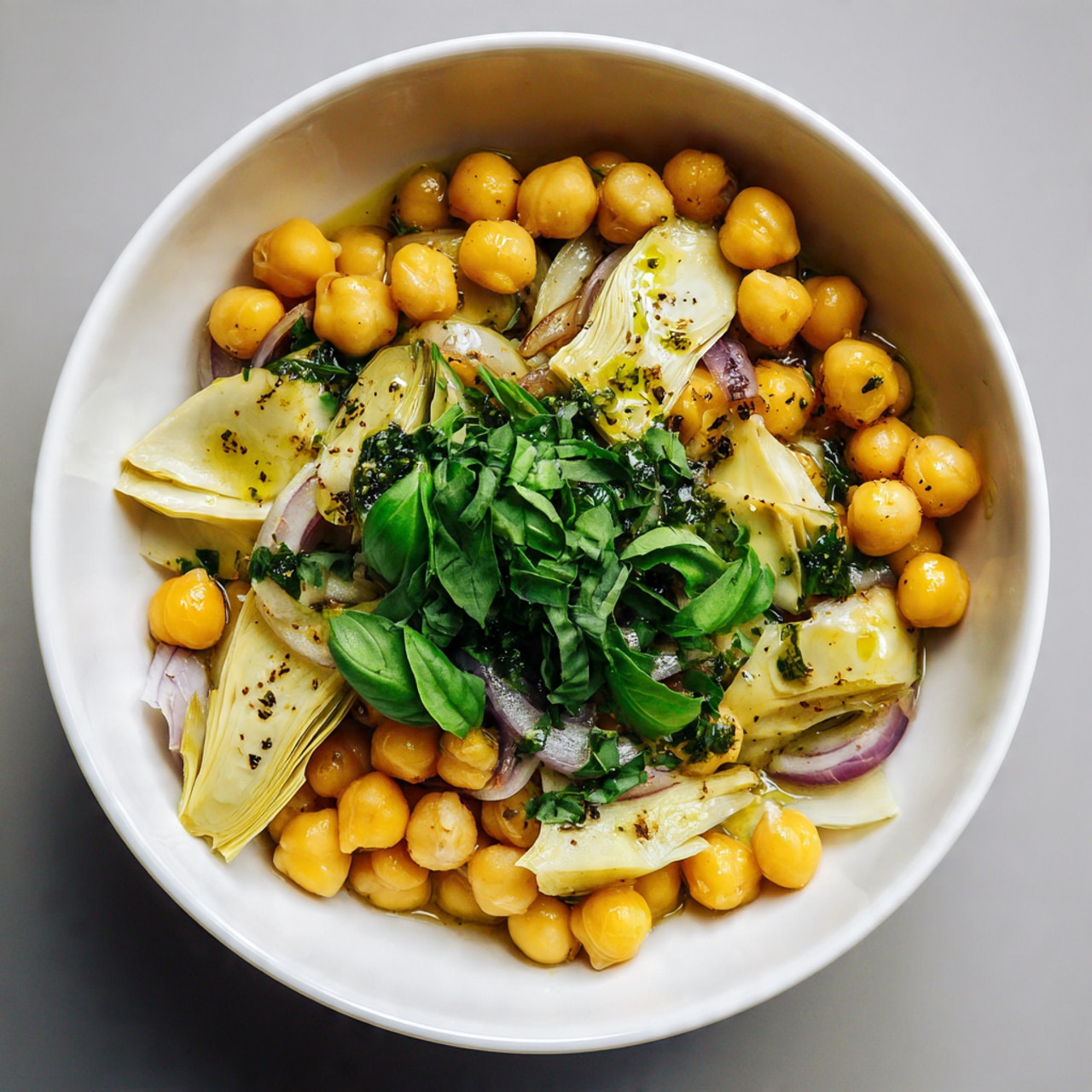 A round white bowl sits on a white marbled surface, filled with layers of bright yellow chickpeas that cover most of the bowl. Mixed with the chickpeas are pieces of pale yellow artichoke hearts placed mainly on the sides and center. Thin slices of light purple onion are partly visible under the chickpeas around the edges. On top, there is a pile of fresh green chopped basil and herbs, with a drizzle of shiny green olive oil and flecks of dark seasoning scattered all over. The colors are fresh and natural, with the yellow, green, and purple contrasting nicely against the white bowl photo taken with an iphone --ar 4:5 --v 7