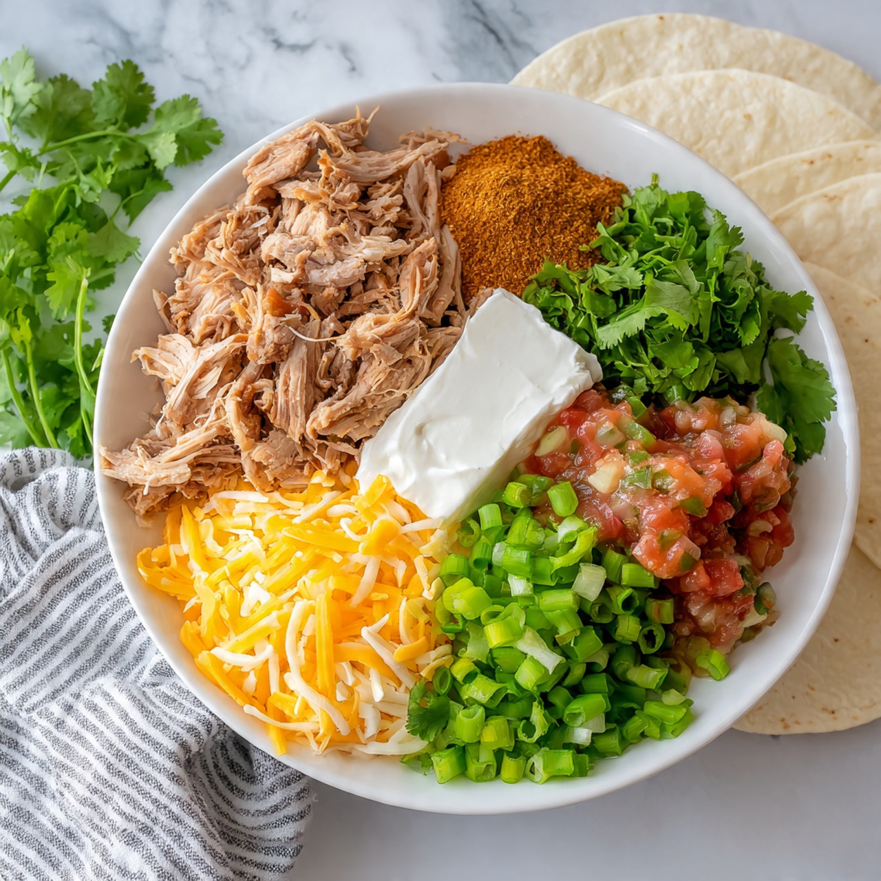 A white bowl on a white marbled surface holds several separate ingredients arranged in a circular layout. Starting from the top left and moving clockwise, there is a pile of light brown shredded cooked chicken, an orange-brown heap of seasoning powder, a bunch of bright green fresh cilantro leaves, a mound of shredded yellow and white cheese, a large white block of cream cheese, a small pile of chopped bright green onions, and a scoop of chunky red tomato salsa with bits of green peppers. A striped gray and white cloth is placed to the left side of the bowl, and two white tortillas rest on the right side of the frame. photo taken with an iphone --ar 4:5 --v 7