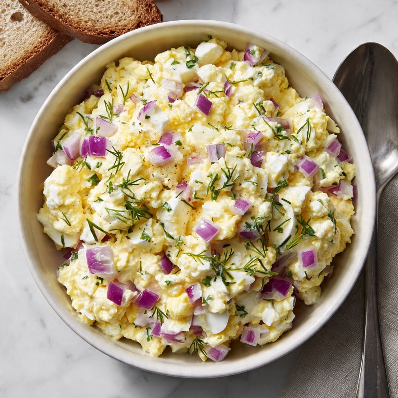A white bowl filled with a creamy egg salad showing about three layers of ingredients mixed together. The base layer is made of chopped hard-boiled eggs with smooth, soft textures in off-white and yellow tones. Throughout the salad, small cubes of bright purple-red onion add a fine texture and two-color contrast. There are also thin green herbs, likely chives or dill, scattered on top and mixed in, creating small green spots over the pale yellow mayo-coated eggs. The bowl sits on a white marbled surface with a piece of light brown bread partially visible on the top left side and a silver spoon on the bottom right. photo taken with an iphone --ar 4:5 --v 7