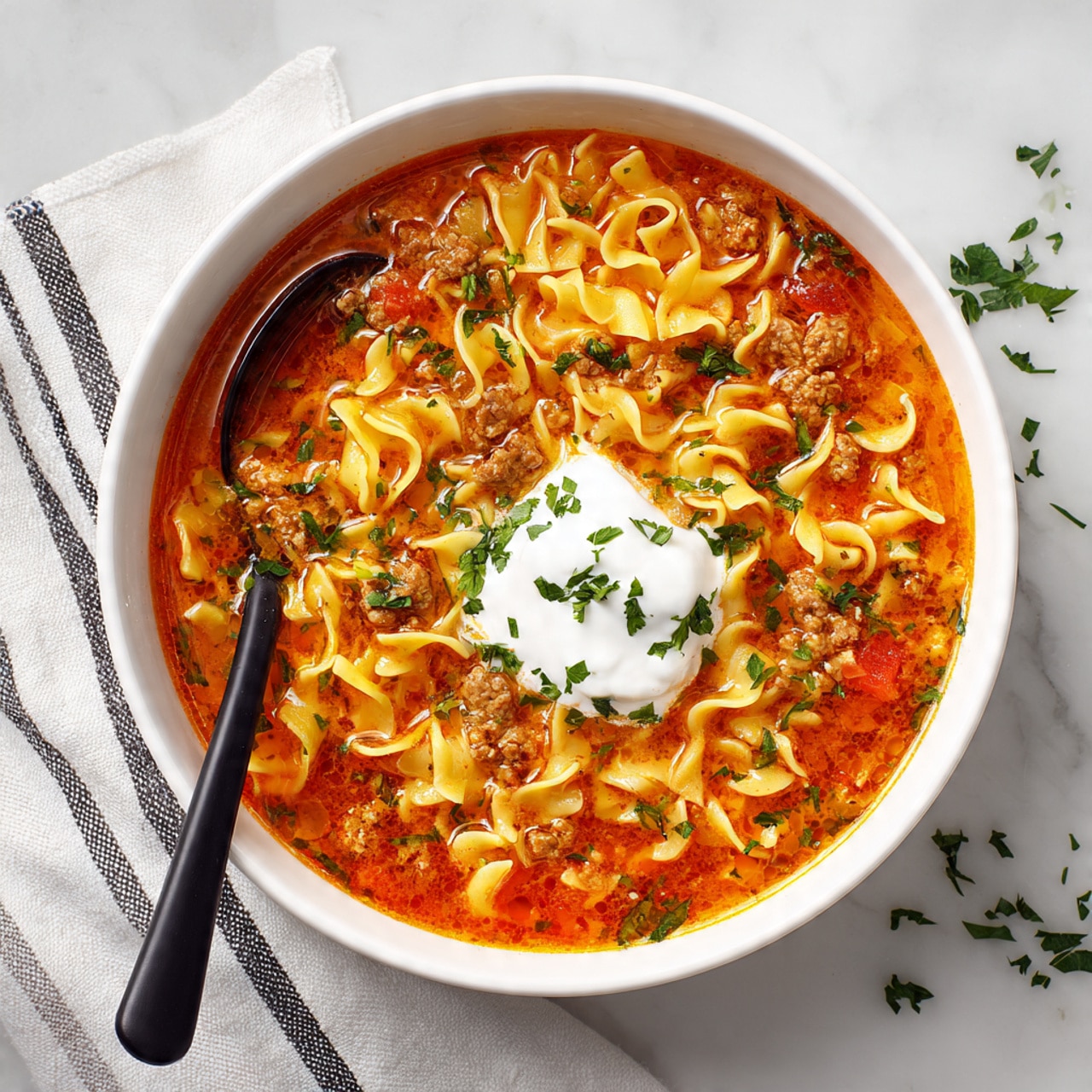 In a white bowl placed on a white marbled surface, there is a rich soup with three main layers: thick orange-brown broth as the base, chunks of browned meat and curved light yellow egg noodles mixed evenly throughout, and a dollop of white sour cream in the center topped with small green herb pieces. A black spoon is partly inside the soup on the left side, with its handle extending outward resting on a white cloth with black stripes. Some green herbs are scattered around the bowl on the surface. photo taken with an iphone --ar 4:5 --v 7