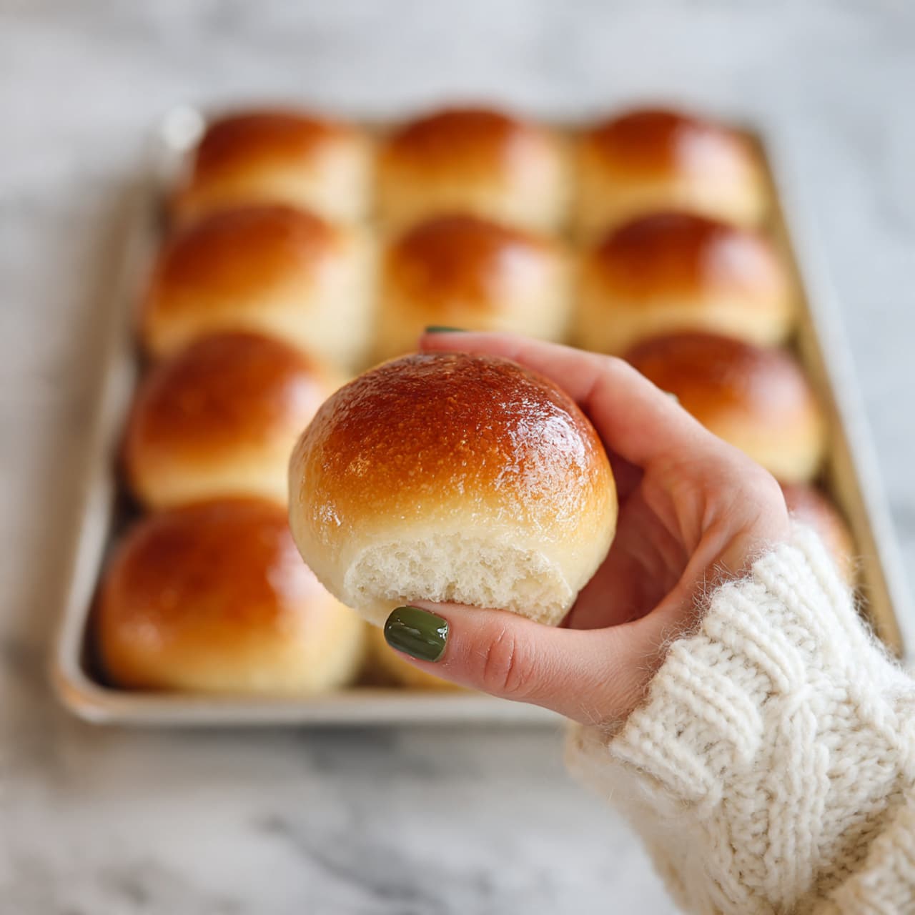 A close-up image shows a woman's hand holding a single soft bread roll with a light brown crust and fluffy, airy inside. In the background, there is a metal tray with twelve similar evenly browned rolls packed closely together. The surface under the tray is a white marbled texture. The bread rolls have a smooth and shiny top crust, each one rounded and uniform in shape. The woman's hand, showing green nail polish and a cozy white sweater sleeve, gently holds the bread roll in the foreground. Photo taken with an iphone --ar 4:5 --v 7