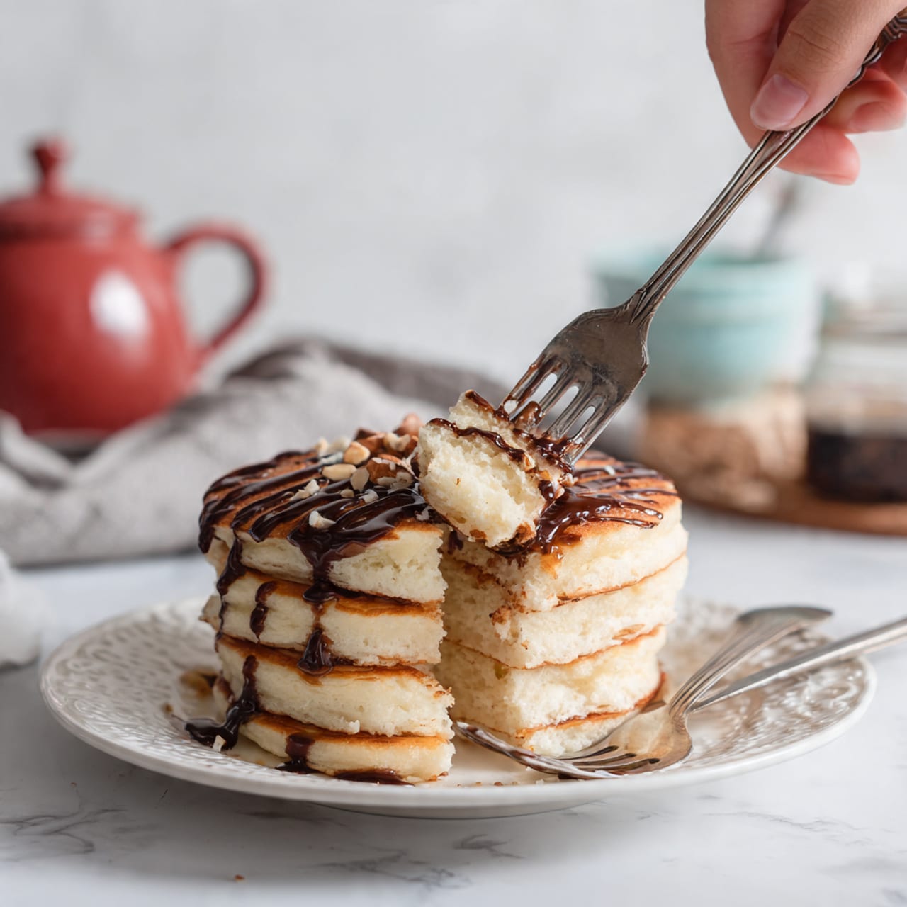 A stack of four thick, light brown pancakes with a soft and fluffy texture sits on a white plate with a decorative pattern, placed on a white marbled surface. The top pancake is drizzled with dark chocolate sauce in stripes and scattered with small almond pieces. A fork and knife rest on the plate, with a woman's hand holding the fork lifting a bite-sized piece from the stack, showing the layers clearly. In the background, there is a blurred red teapot, a bowl, and some glass jars, all on the white marbled surface. photo taken with an iphone --ar 4:5 --v 7