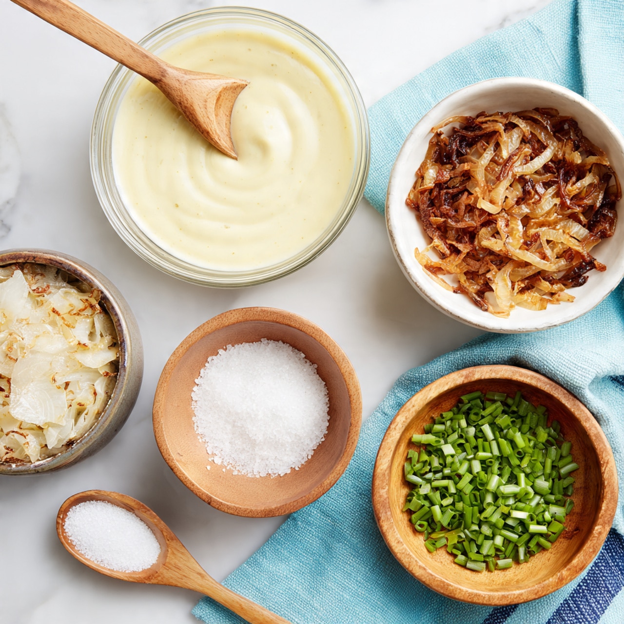 A top view of five containers arranged on a white marbled surface holding different ingredients: a glass bowl with smooth, creamy pale yellow sauce with a wooden spoon resting inside it at the upper left; a white bowl with caramelized, soft brown onions and a wooden spoon inside at the upper right; a small wooden bowl with a swirl of coarse white salt at the middle left; a small wooden bowl with finely chopped bright green chives at the bottom right, placed on a light blue cloth with dark blue lines; and a small wooden spoon holding a small amount of fine white powder at the bottom left. Photo taken with an iphone --ar 4:5 --v 7