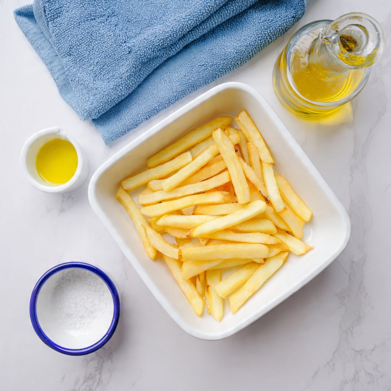 A white rectangular dish holds raw potato fries arranged loosely in one layer, showing a mix of light yellow inner potato flesh with brown skin edges. To the right, there is a small glass bottle with a yellow liquid, likely oil, and to the bottom left, a small white cup with a blue rim containing salt, all placed on a white marbled surface with a blue cloth nearby. The scene is brightly lit, highlighting the fresh texture and natural color of the potato fries photo taken with an iphone --ar 4:5 --v 7