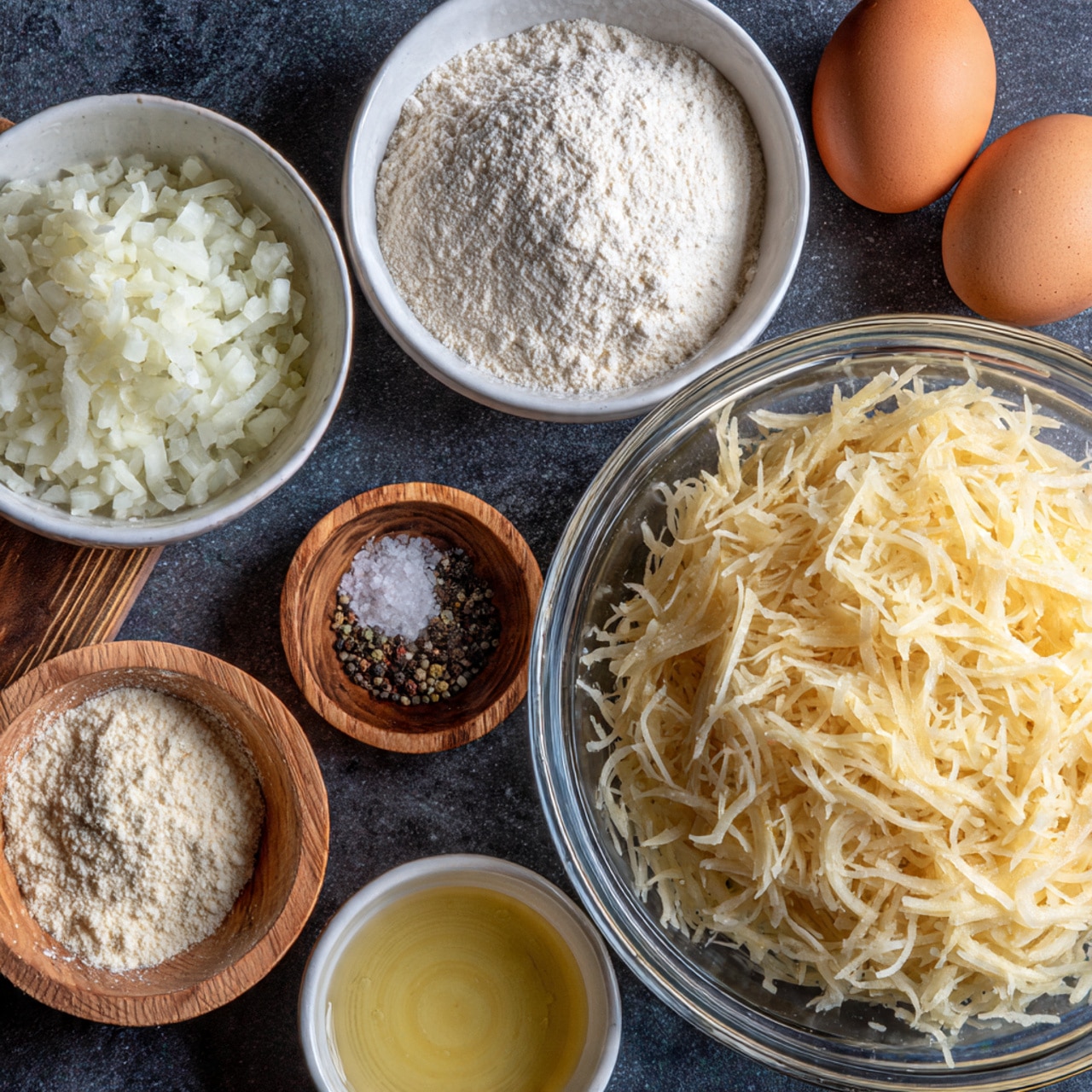 A clear glass bowl on the right filled with a large pile of pale, finely shredded potatoes. To its left, smaller white bowls hold different ingredients: the top white bowl contains finely chopped white onions, the bottom white bowl has a mound of white flour, and a small wooden bowl holds white salt and black pepper. Two brown eggs are placed between the bowls and a small white bowl with a light yellow liquid, all set on a dark, smooth surface. photo taken with an iphone --ar 4:5 --v 7