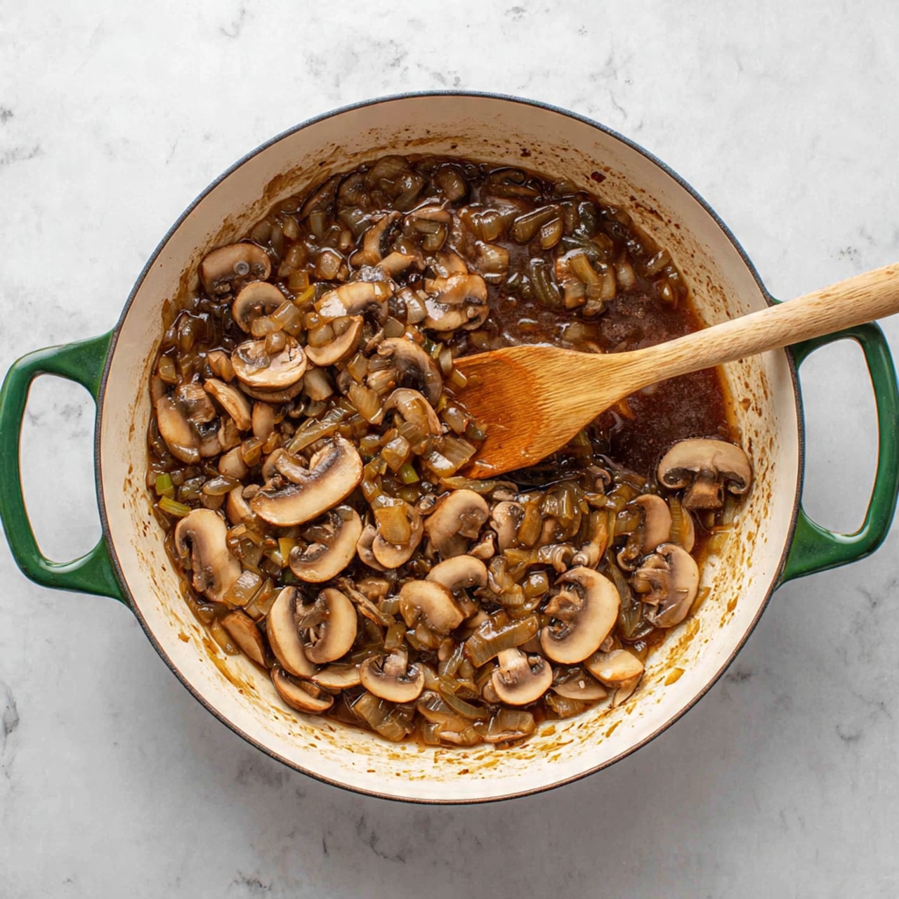 A white pan with green handles filled with cooked mushrooms and onions in a brown sauce. The mushrooms are sliced and sit mostly on one side of the pan, showing some browned bits on the white pan surface. A light wooden spoon is resting in the pan, mixing the mushrooms and onions. The background is a white marbled texture. photo taken with an iphone --ar 4:5 --v 7