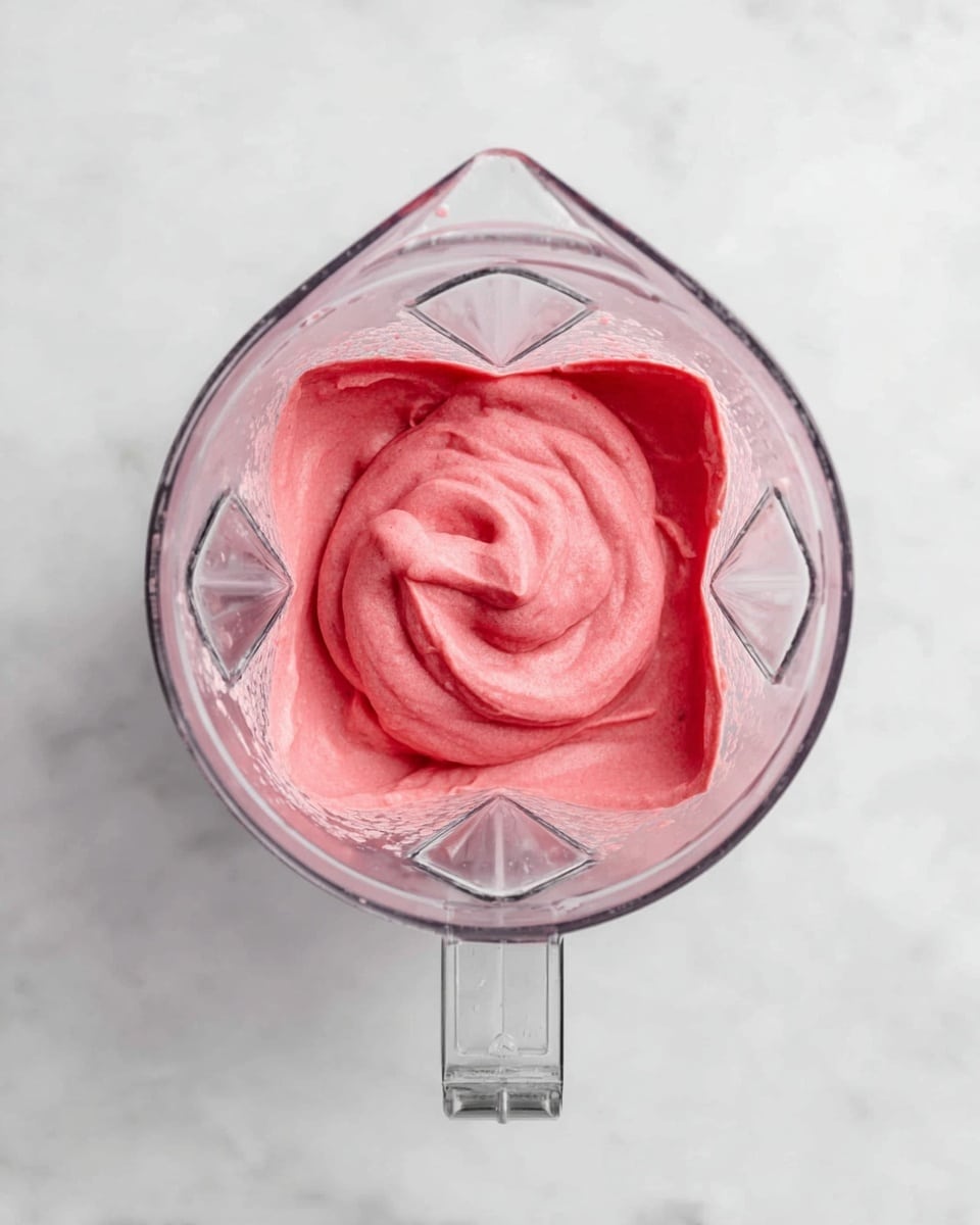 A clear blender pitcher with a square inner container holds a smooth, thick pink puree swirled gently on top in soft peaks. The texture looks creamy and dense, with tiny air bubbles giving a light, fluffy look. The blender sits on a white marbled surface, adding a clean, bright contrast to the rich pink color. Photo taken with an iphone --ar 4:5 --v 7