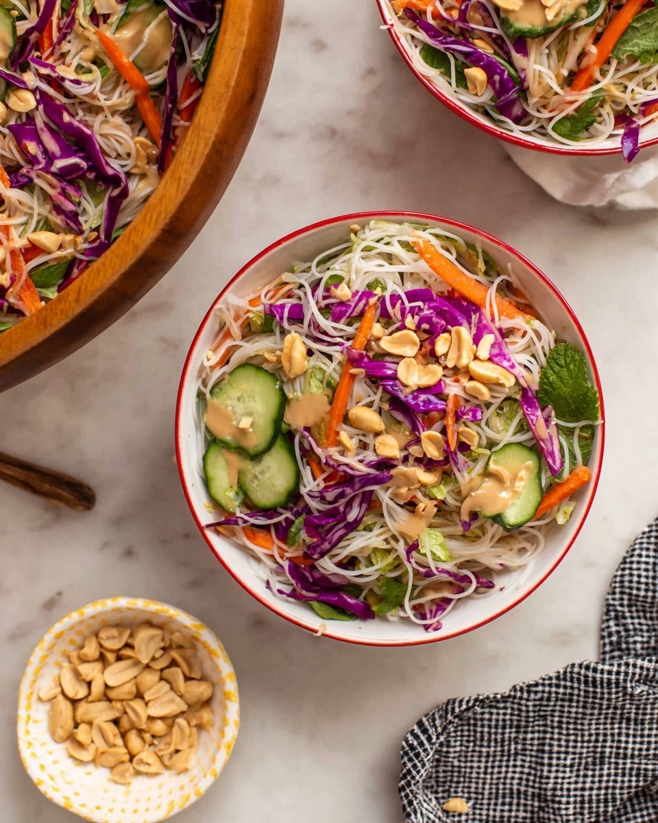The image shows a fresh noodle salad served in two white bowls with thin red rims, placed on a white marbled surface. The salad has a base layer of thin white noodles, topped with bright purple shredded cabbage and thin orange carrot strips scattered on top. There are several green cucumber slices laid on the salad, and light brown peanuts sprinkled over everything. Some fresh green herbs are mixed throughout and a drizzle of light beige peanut sauce sits on the noodles. On the left side, part of a large wooden bowl filled with the same noodle salad is visible. A small white dish with yellow trim holding peanuts is in the lower left corner, and a black and white checkered cloth peeks into the frame. Photo taken with an iphone --ar 4:5 --v 7