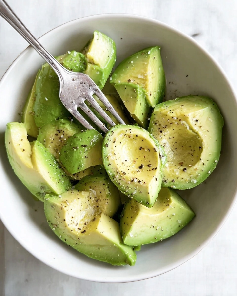 In a white bowl, there are several avocado halves and chunks arranged close together, showing a mix of smooth light green inside and slightly darker green outer edges. The avocado pieces are sprinkled with black pepper and a bit of salt, creating small black and white dots all over. A silver fork rests on top of the avocados, with the tines slightly pressed into one avocado chunk. The bowl sits on a white marbled surface, adding a clean and simple look to the scene. Photo taken with an iphone --ar 4:5 --v 7