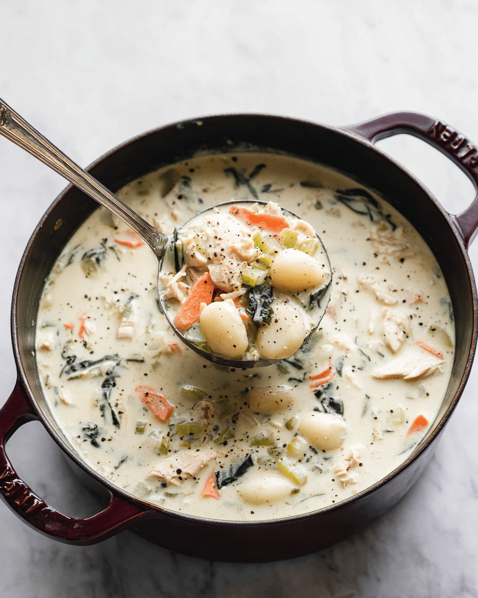A round dark red cast iron pot filled with thick creamy white soup containing visible pieces of light pink chicken, pale green celery, small orange carrot strips, dark green leafy herbs, and white gnocchi-like shapes. The soup has a smooth, rich texture with some black pepper sprinkled on top. A silver ladle with a long handle is lifting a portion of the soup with chunks of chicken, celery, carrot, and gnocchi from the pot. The pot is placed on a white marbled surface. photo taken with an iphone --ar 4:5 --v 7