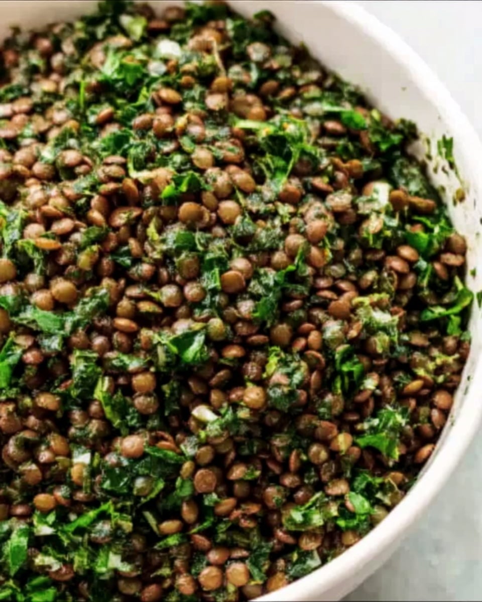 A close-up view of a white bowl filled with a mix of small brown lentils and finely chopped dark green leafy herbs. The lentils appear soft and slightly shiny, contrasting with the bright, fresh texture of the chopped greens. The bowl sits on a white marbled surface, with the mixture evenly spread inside, showing a simple, fresh dish with natural colors and textures. Photo taken with an iphone --ar 4:5 --v 7