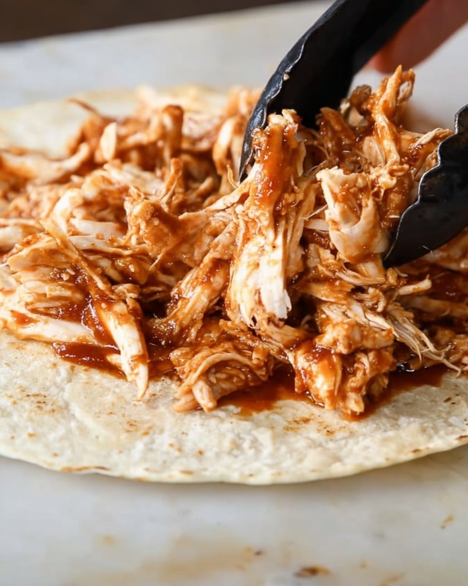 A close-up view of a single crispy, light beige tortilla placed flat on a white marbled surface, topped with a generous layer of shredded cooked chicken covered in a rich reddish-brown sauce. The chicken has a moist texture with visible strands and small chunks mixed together. A woman's hand is seen on the right side holding black tongs, gently lifting some shredded chicken. The background remains soft and out of focus, emphasizing the tortilla and chicken. photo taken with an iphone --ar 4:5 --v 7
