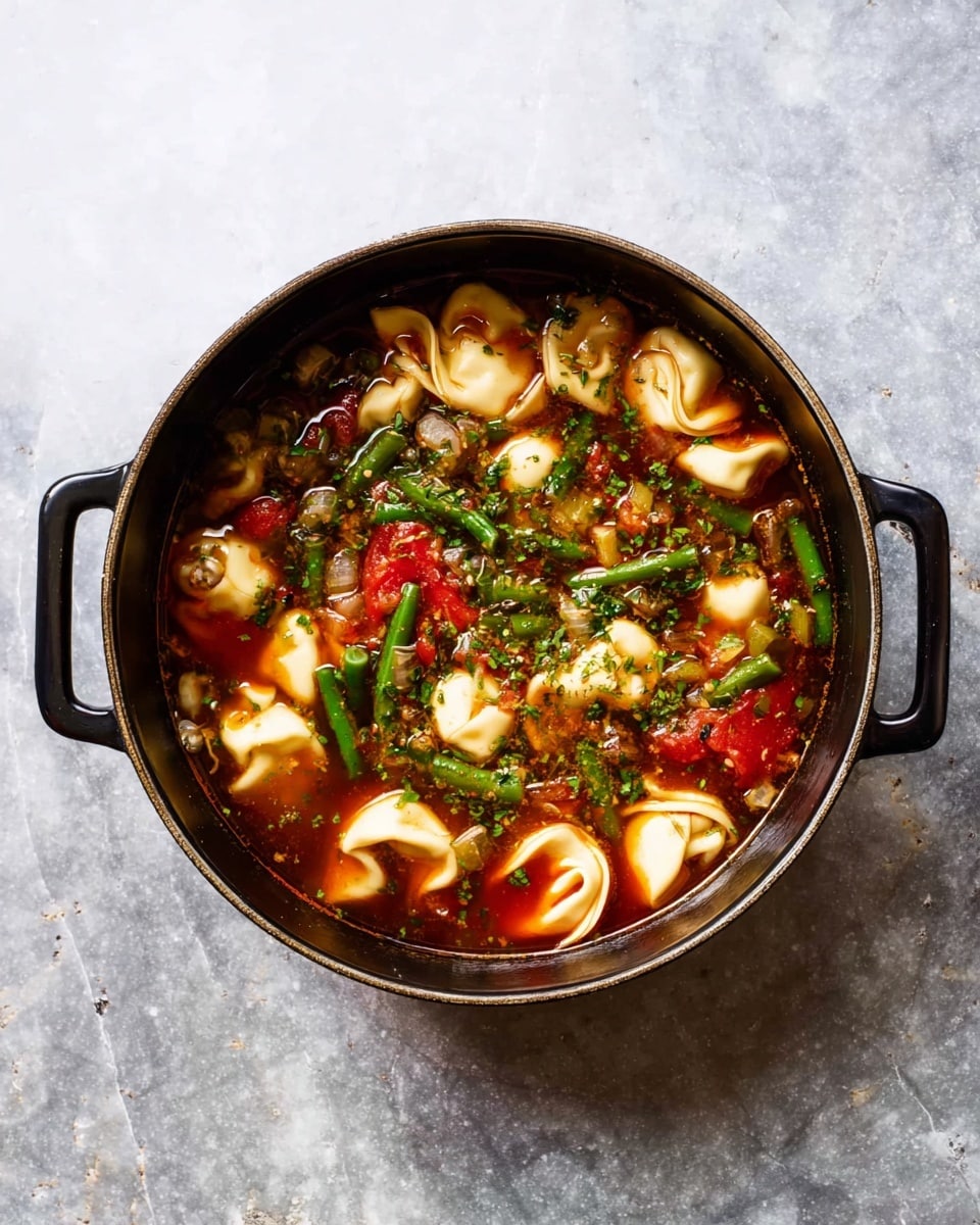A black round pot with two handles holds a rich soup with three main visible layers: the bottom layer is a dark broth, the middle layer has bright red tomato pieces mixed with chopped green beans and other small vegetables, and the top layer features white tortellini pasta floating throughout. Tiny bits of herbs sprinkle the top, adding texture, all placed on a white marbled surface. photo taken with an iphone --ar 4:5 --v 7
