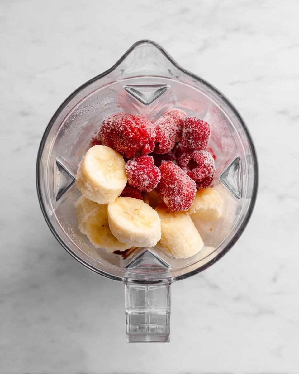 A clear blender container is shown from above, filled with two main layers of frozen fruit. The bottom layer has several thick slices of pale yellow frozen banana with a slightly frosty texture. Above the banana layer, there are whole frozen strawberries that appear bright red with icy crystals on their surface. The blender sits on a white marbled surface. photo taken with an iphone --ar 4:5 --v 7