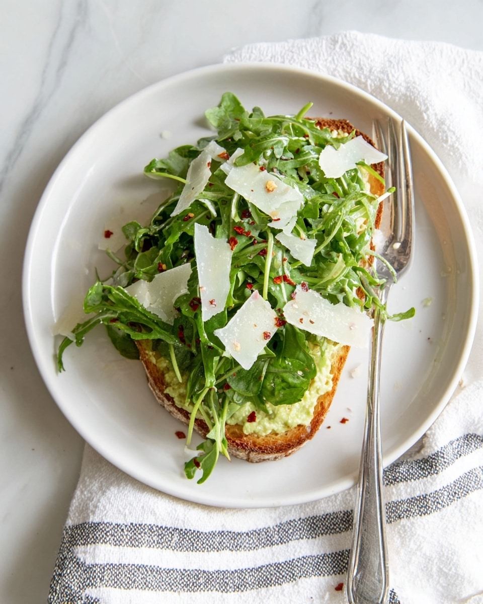 A single piece of light golden toasted bread forms the base layer on a white plate, topped with a creamy pale green spread. Over the spread, fresh bright green arugula leaves are scattered thickly, adding a leafy texture. Thin, off-white shavings of cheese are placed on top of the arugula, with small red specks sprinkled across the whole dish. A metal fork rests on the right edge of the plate, and the plate is set on a white marbled surface with a white cloth that has thin, dark stripes underneath. Photo taken with an iphone --ar 4:5 --v 7