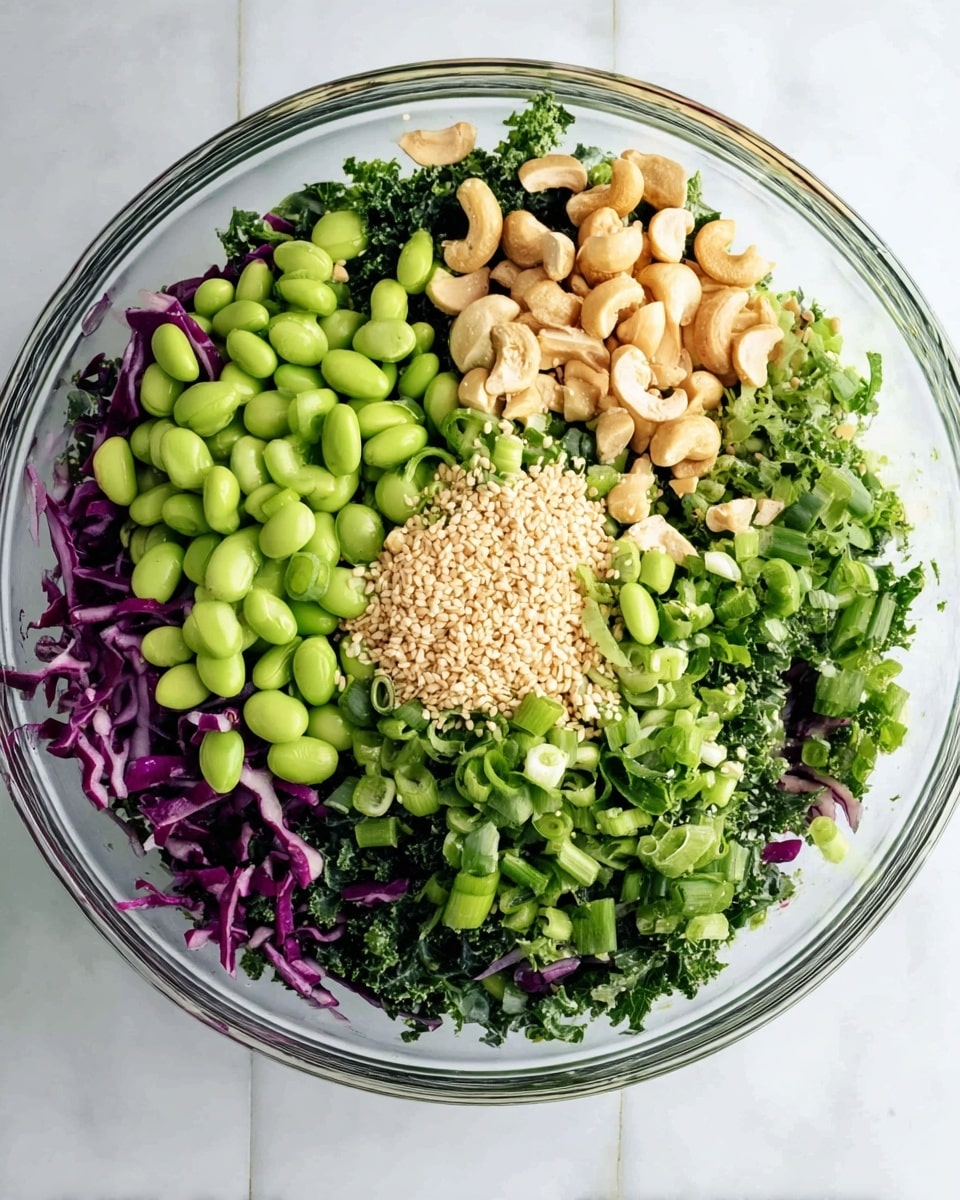 A clear glass bowl filled with layers of colorful salad ingredients sits on a white marbled surface. The bottom layer is chopped dark green kale mixed with shredded purple cabbage, creating a vibrant base. On top, bright green edamame beans cover about a third of the bowl’s left side. The center has a small pile of light beige sesame seeds surrounded by chopped light green scallions. To the right, there are roughly chopped cashew nuts and more scallions scattered on top. Fresh green cilantro leaves peek through, adding more green tones. The dish looks fresh, healthy, and full of different textures. Photo taken with an iphone --ar 4:5 --v 7